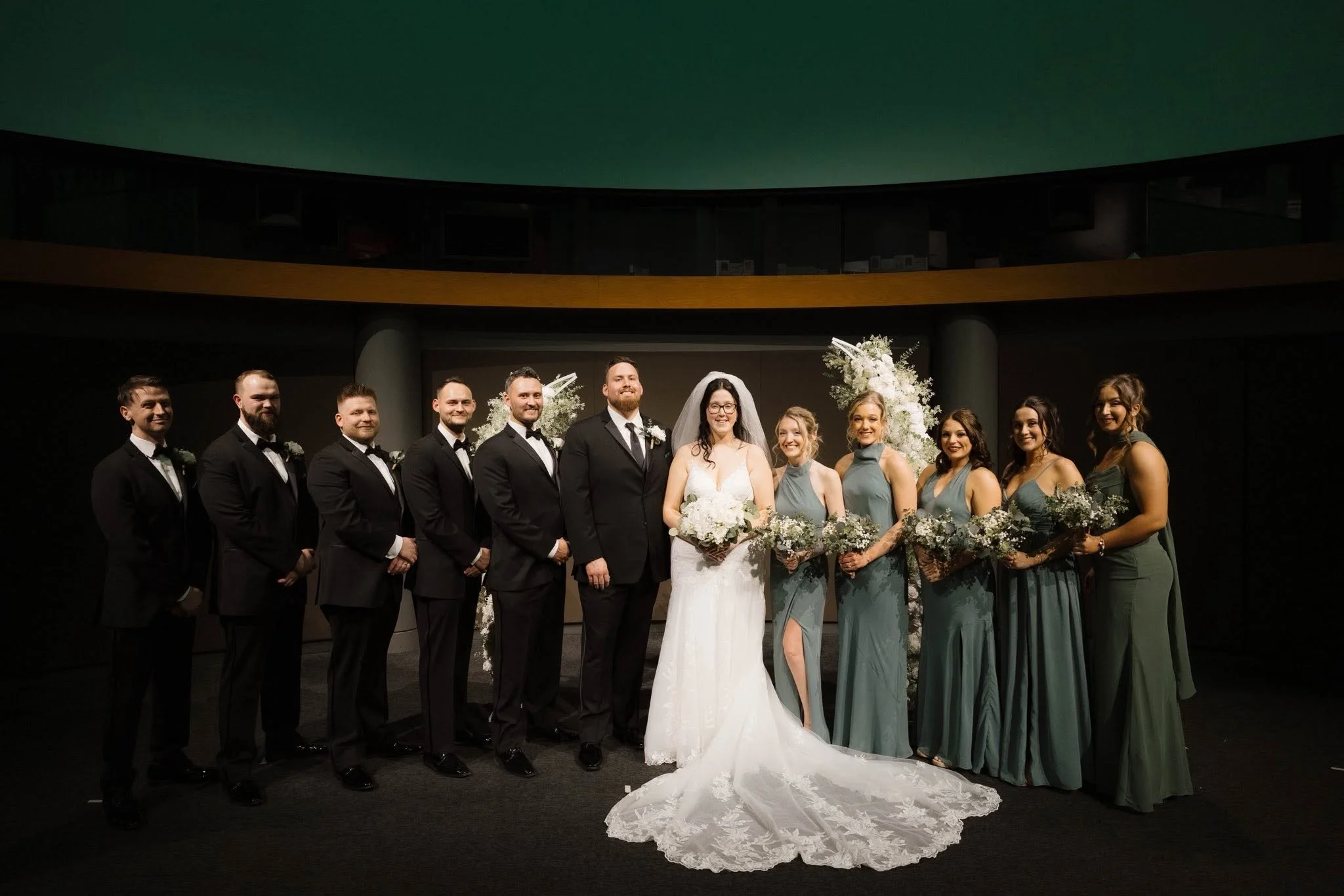 Group of wedding guests, including the bride and groom, standing in a line on a stage with floral decorations, against a dark background.