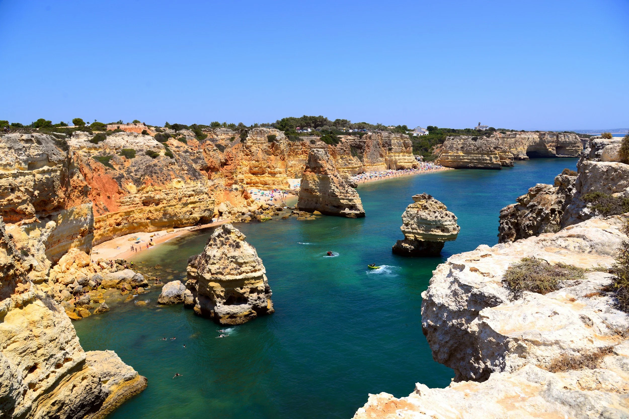 Cliffs along a beach with turquoise water, rocks and people swimming and sunbathing on the sandy shore, clear blue sky