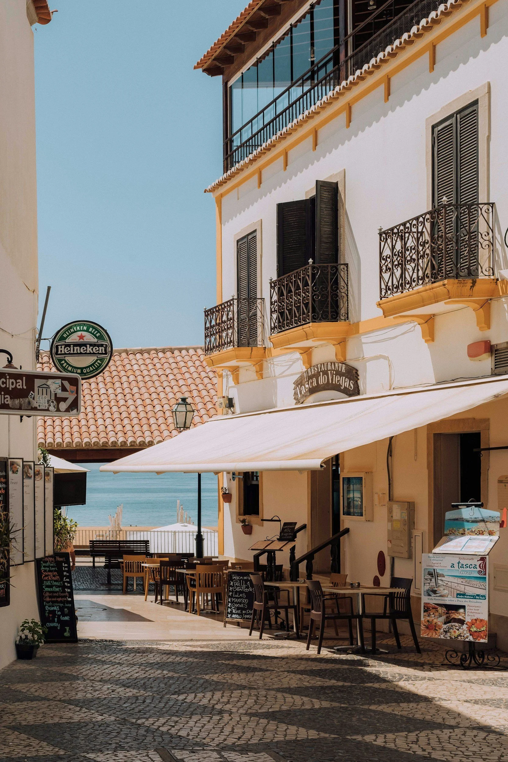 Outdoor cafe with tables and chairs, blackboards with menu, umbrellas, overlooking the ocean under clear skies, with a white and yellow building with black shutters and decorative railings