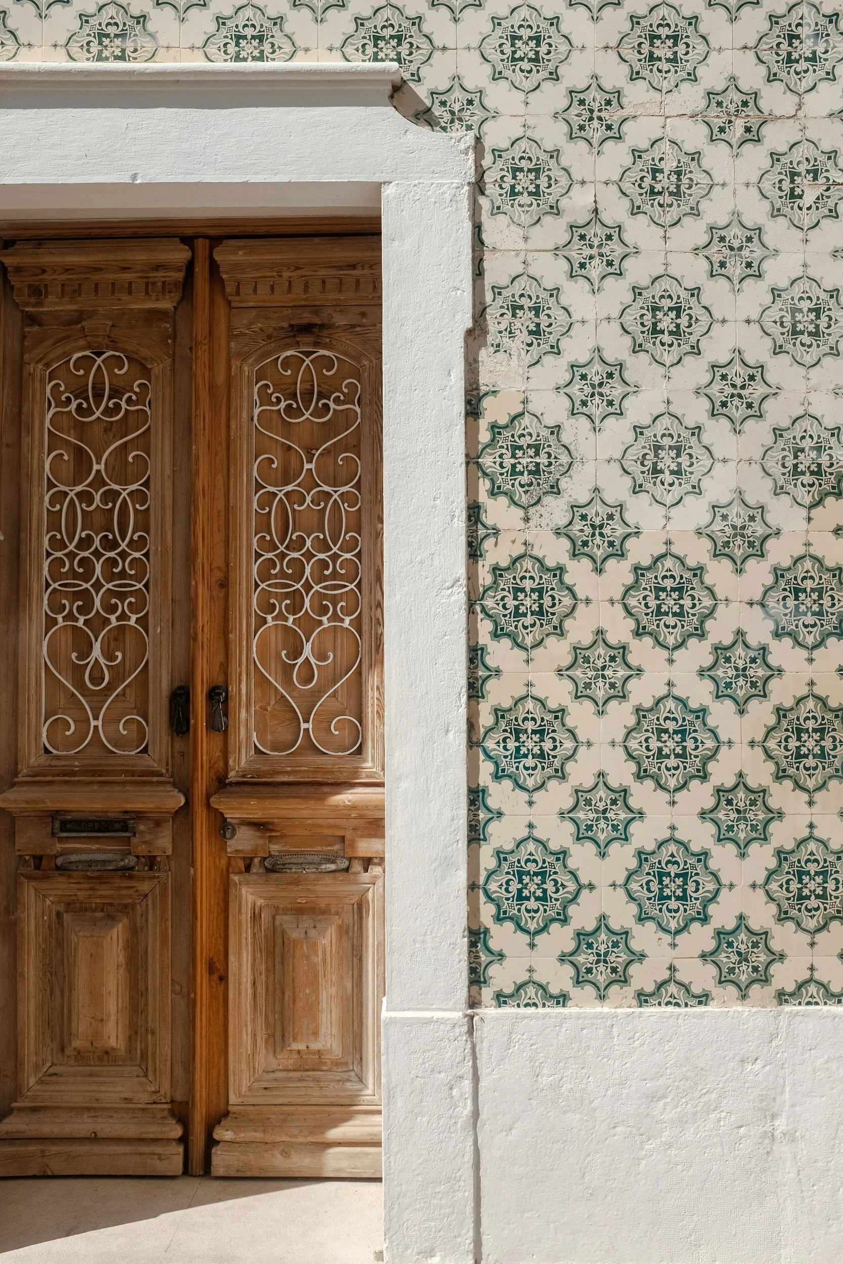 Wooden door with intricate ironwork and mail slots, framed by a white stucco wall, with decorative green and white patterned tiles on the right side.