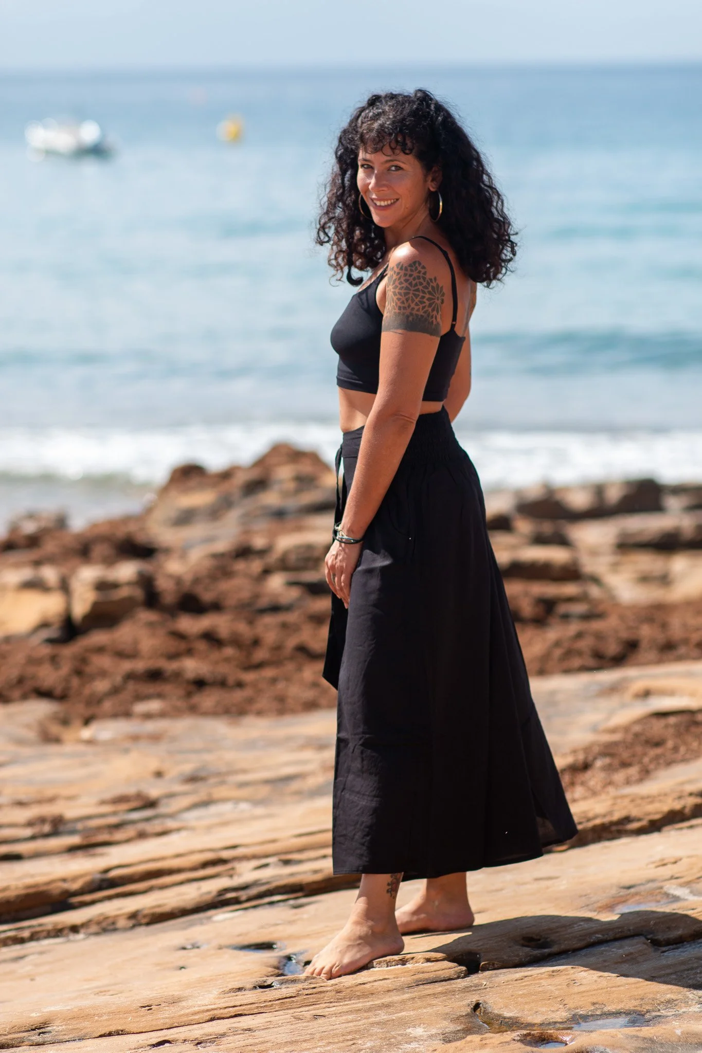 A woman with curly black hair, wearing a black crop top and black skirt, standing barefoot on a rocky beach near the ocean, smiling at the camera.