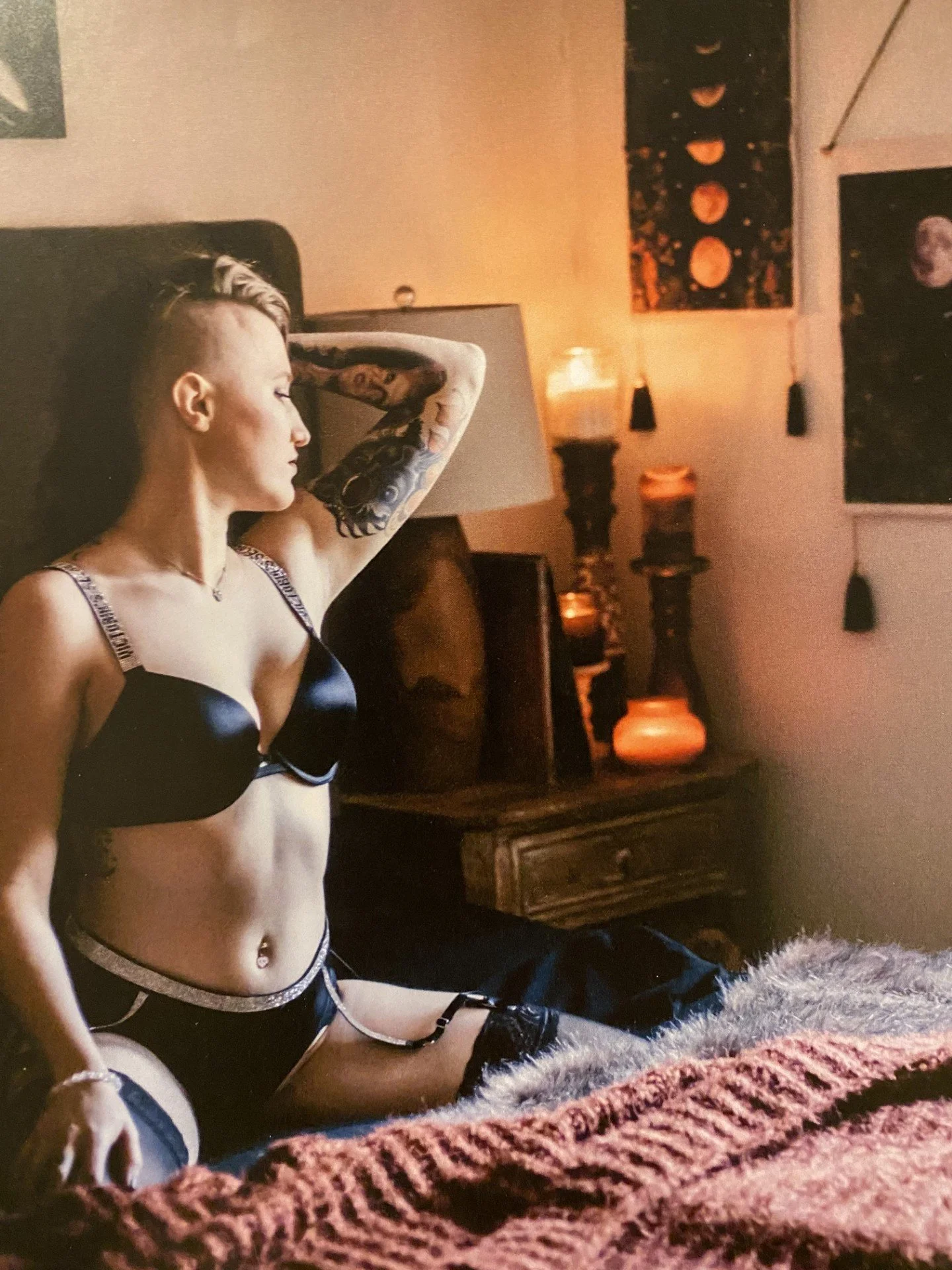A woman with tattooed arms and a short hairstyle, wearing black lingerie with silver accents, sitting on a furry blanket in a dimly lit room with art and candles on the wall.