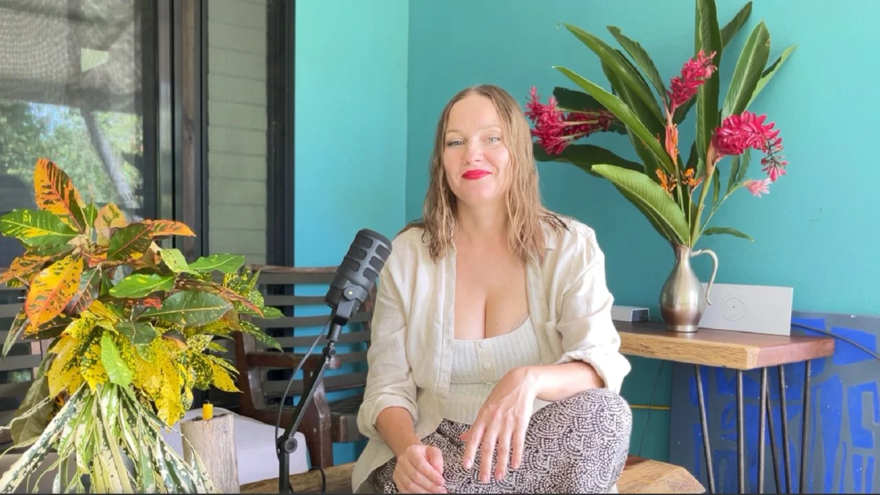 Woman sitting at a table with a microphone, green plants and pink flowers behind her against a teal wall.