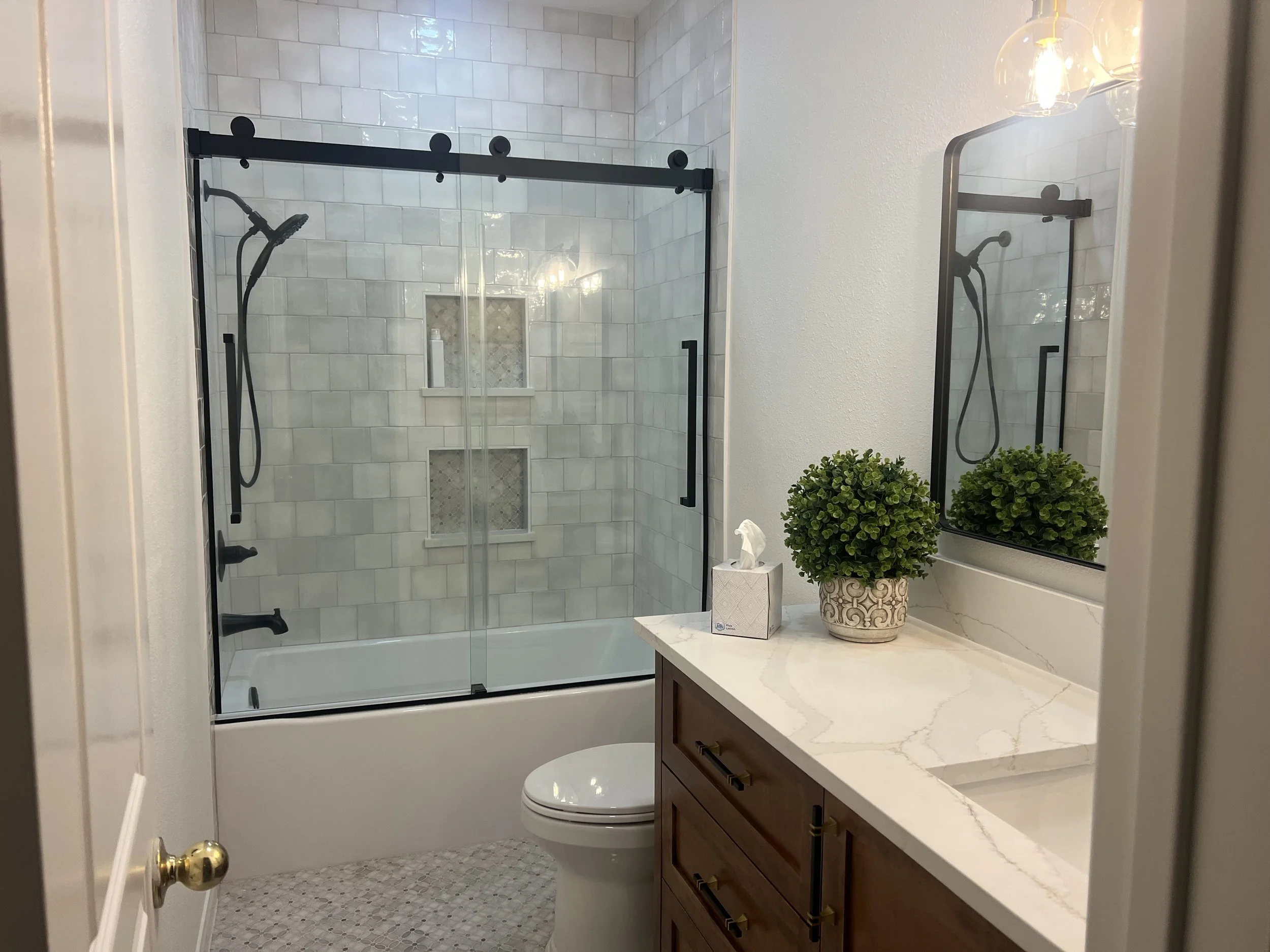 A bathroom with a glass-enclosed shower, white tiled walls, a white toilet, a marble countertop with a wooden cabinet below, a rectangular mirror, a potted plant, and a tissue box on the counter.