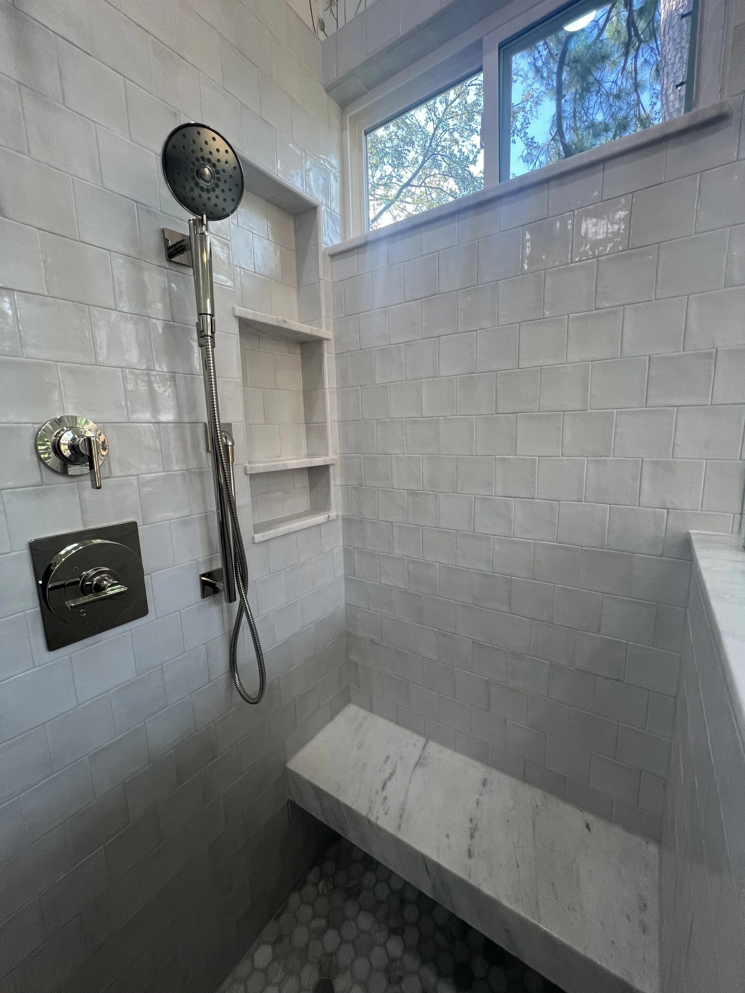 Shower area with white subway tiles, a black showerhead, a built-in niche, a marble bench, and a window showing trees outside.