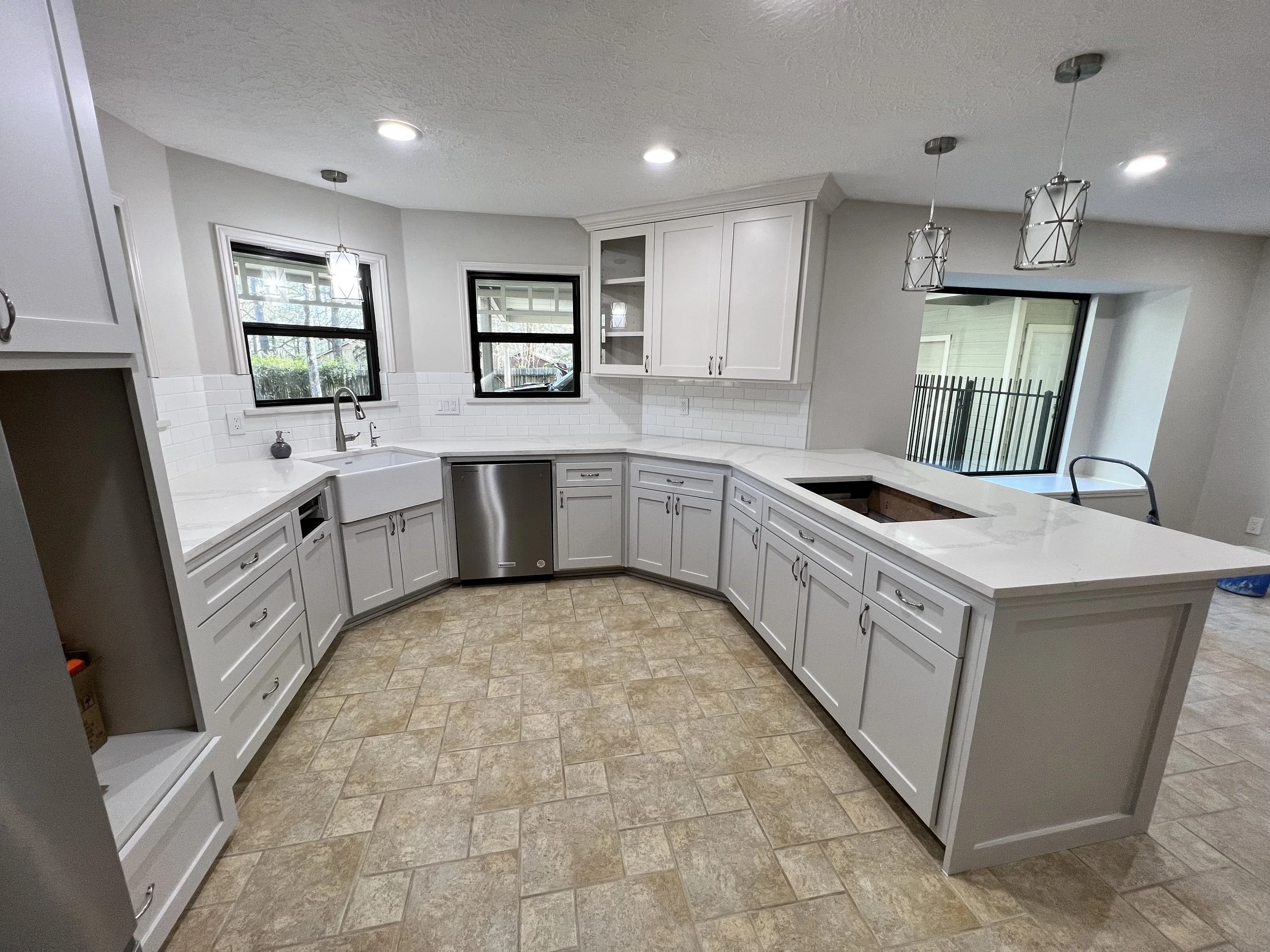 A kitchen with white cabinets, a granite countertop, a white farmhouse sink, two windows, and pendant lights. There is a sliding glass door leading outside and beige tiled floor.