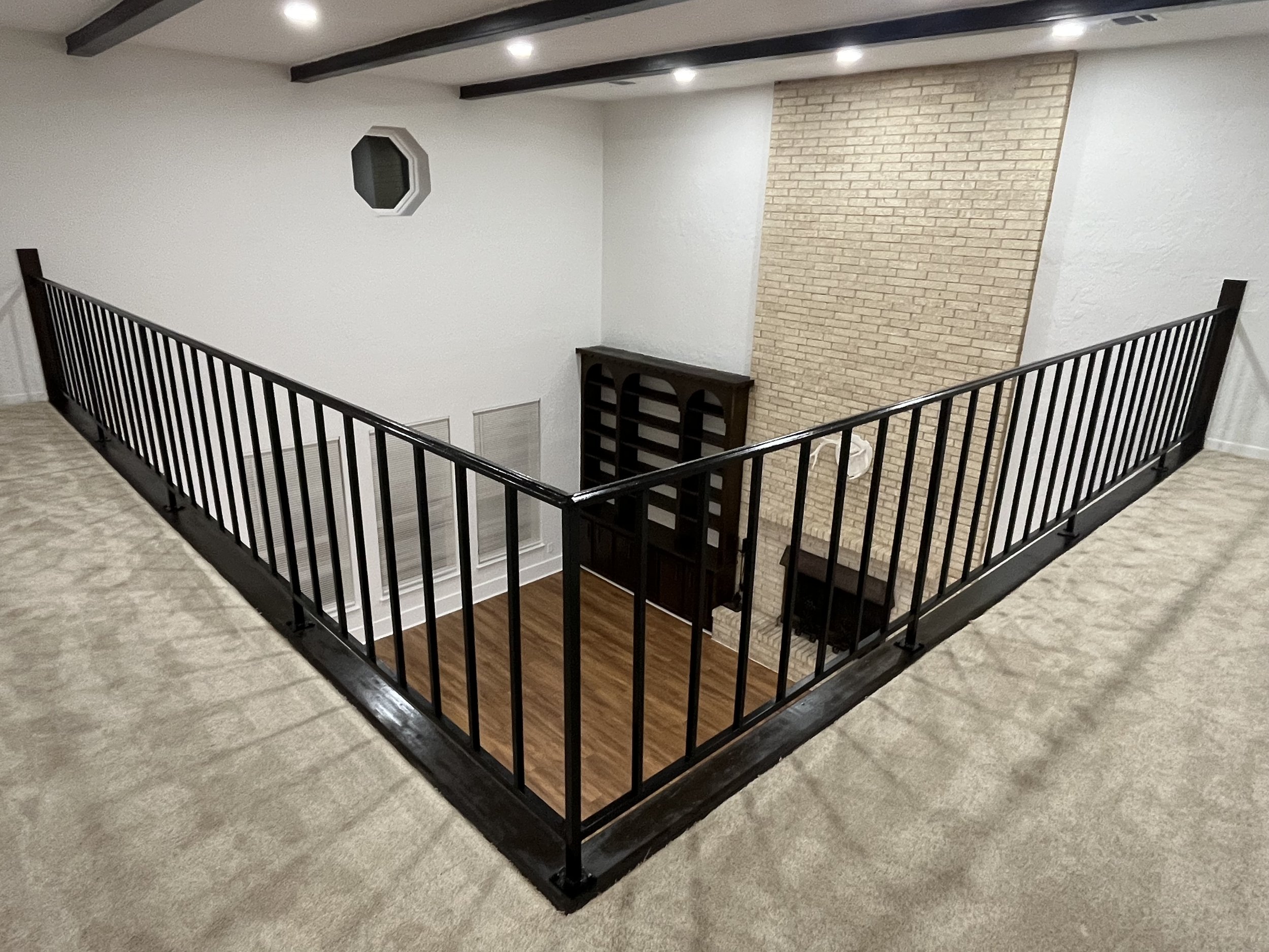A staircase with black railings and a textured brick fireplace on the wall at the top, featuring a small dark wooden shelf underneath the brick section. The floor is light-colored hardwood, and the ceiling has dark wooden beams with recessed lighting