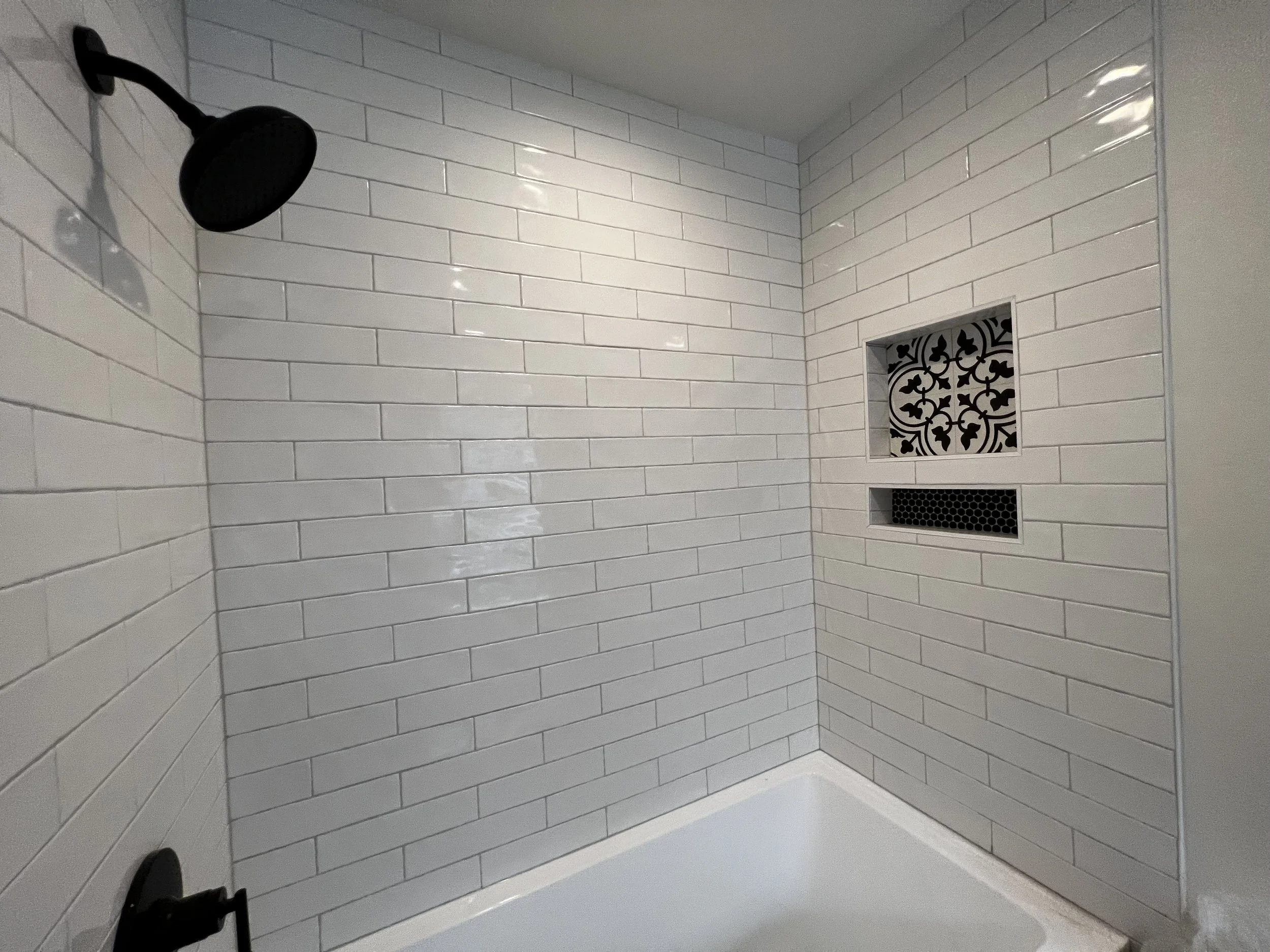 White tiled shower walls with a black showerhead, two built-in shelves, and a small decorative tile accent.