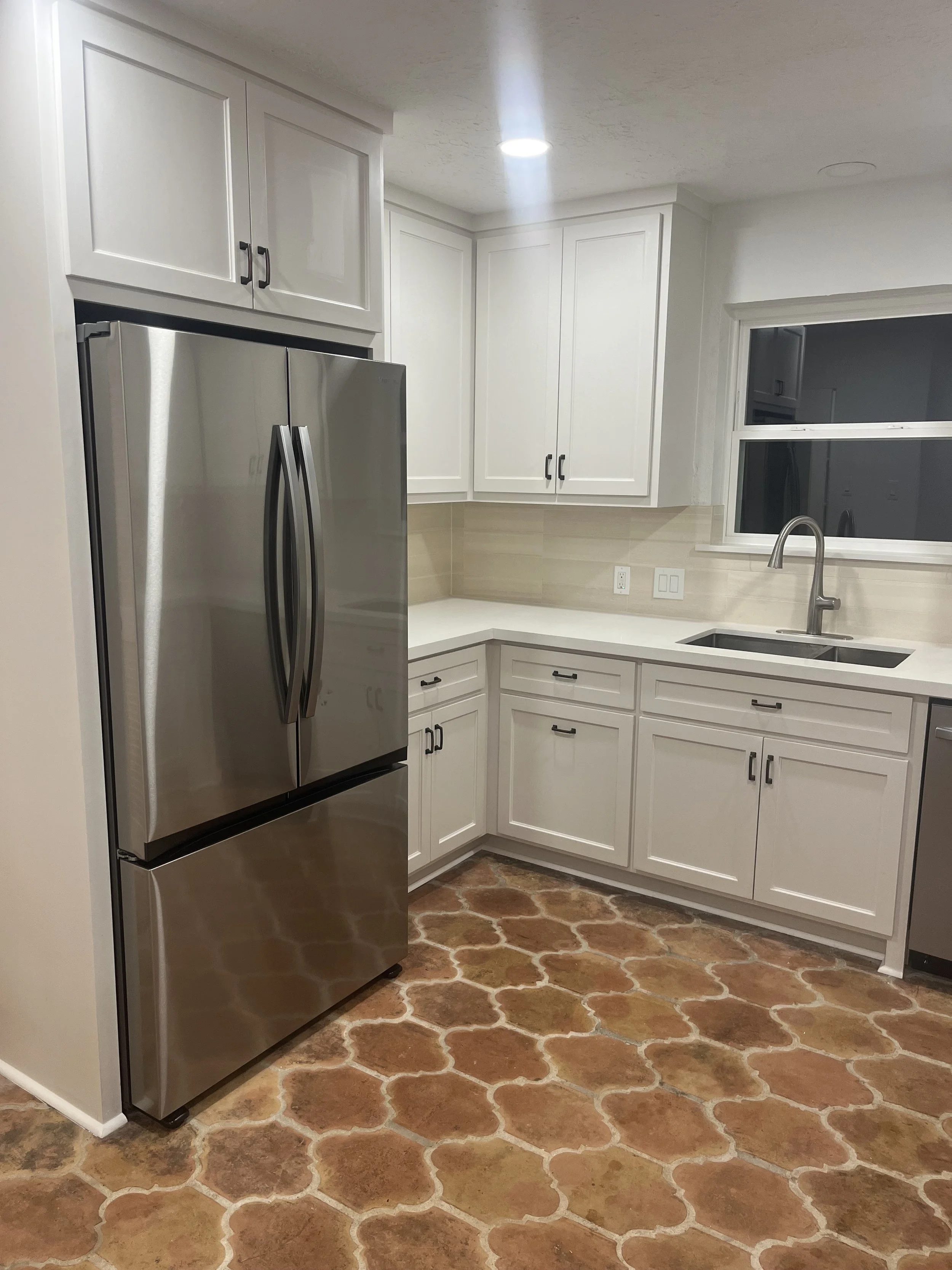 A modern kitchen with white cabinets, a stainless steel refrigerator, a large window above the sink, and a terracotta tile floor with a Moroccan-style pattern.