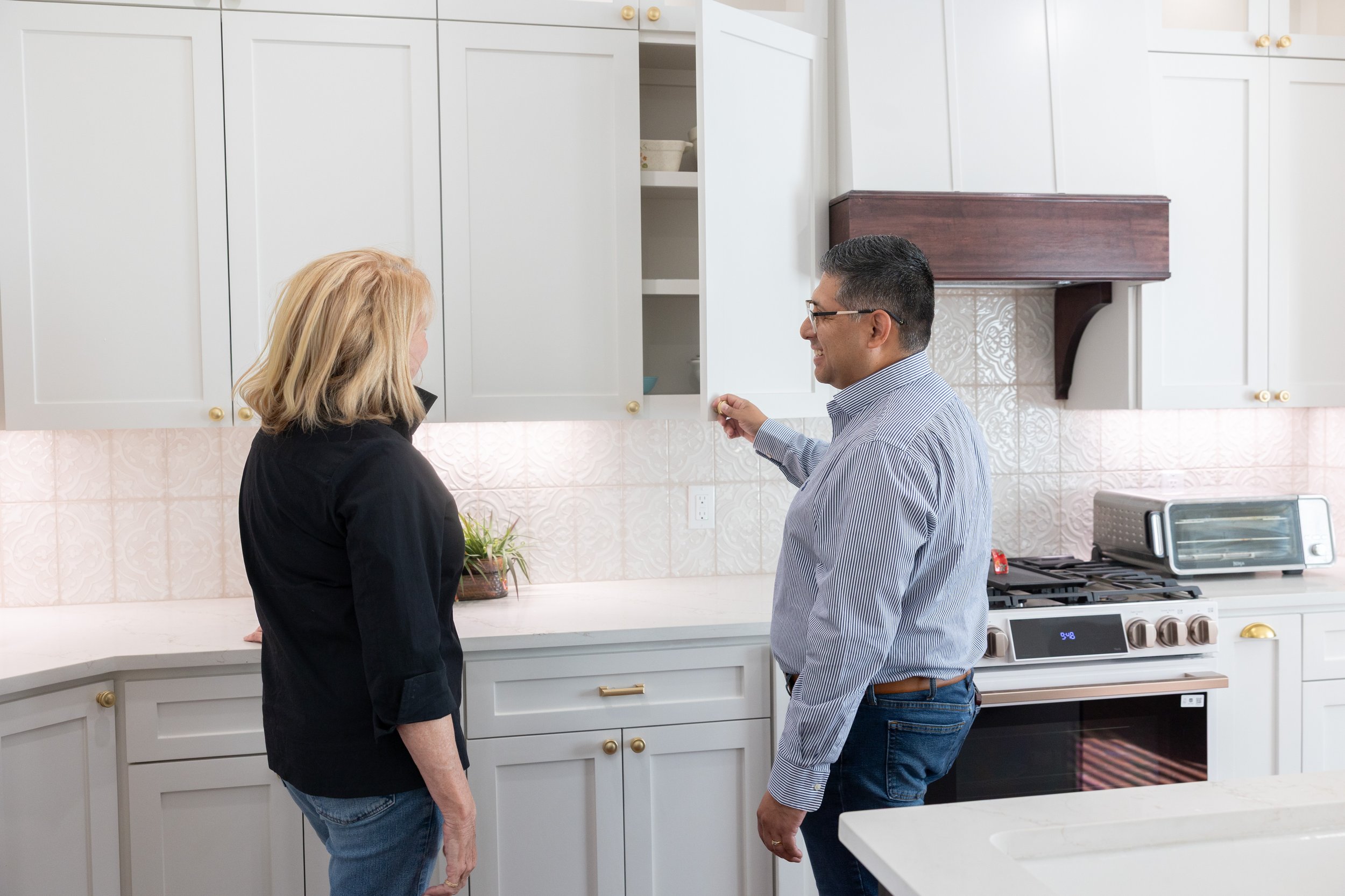 A man and a woman standing in a white kitchen, holding hands and smiling at each other.