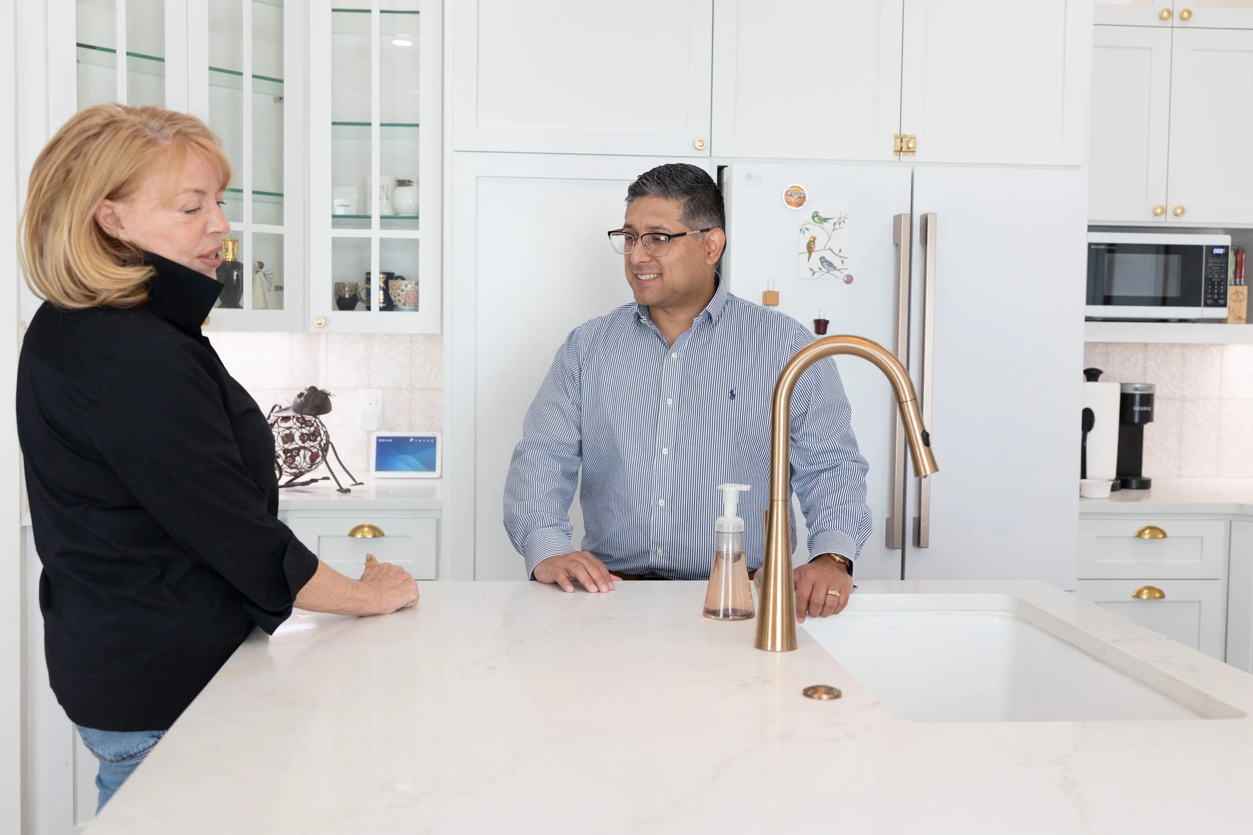 A woman and a man are standing at a kitchen island, shaking hands. The woman has blonde hair and is wearing a black top, while the man has dark hair, glasses, and is wearing a striped shirt. The kitchen has white cabinets, a gold faucet, and various 