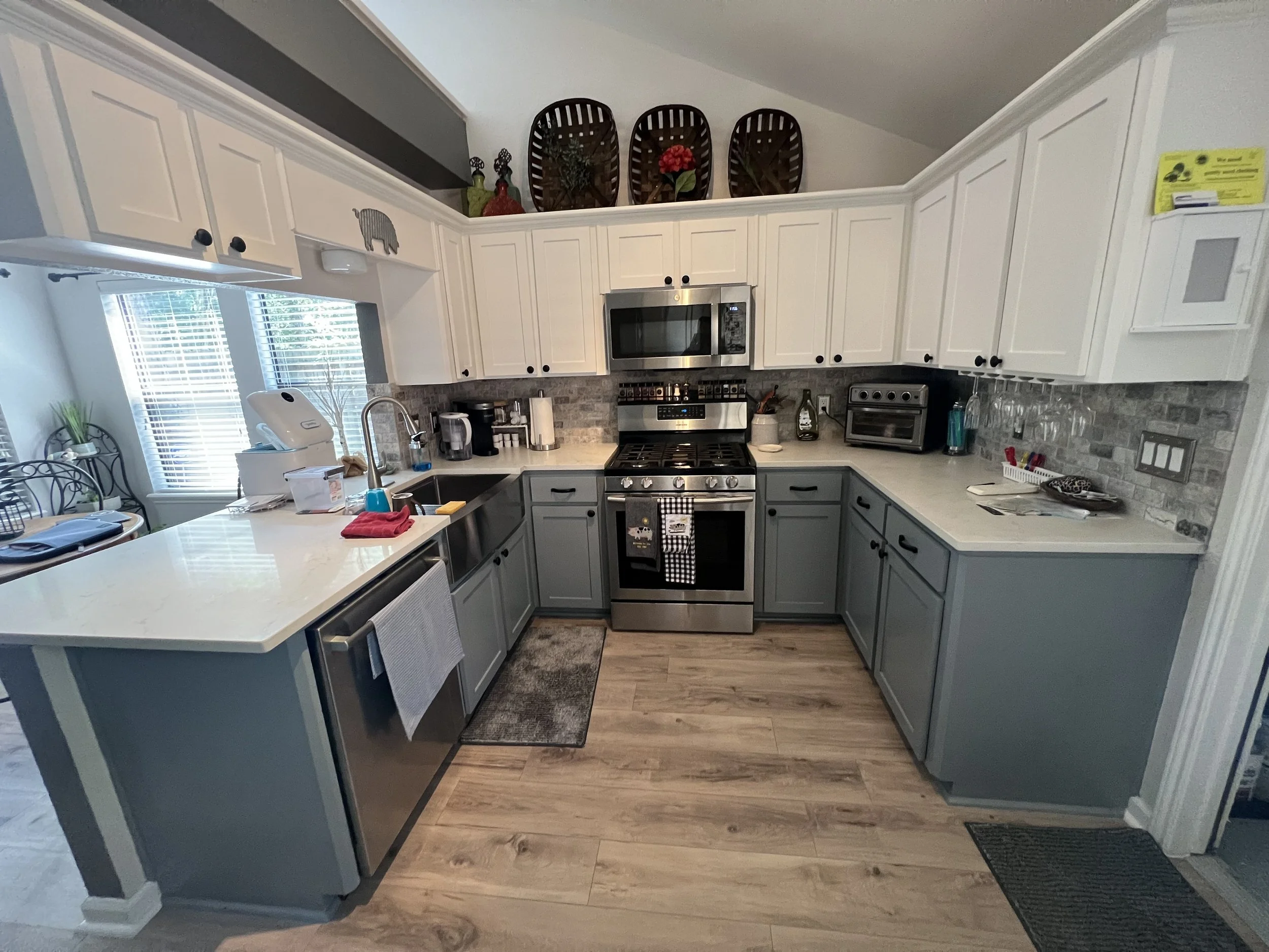 Kitchen with white upper cabinets, gray lower cabinets, stainless steel appliances, black knobs, and a window with blinds. Decor includes green plants, red flowers, and a pig-shaped ornament on the cabinets. Counter has appliances, dish rack, and ute