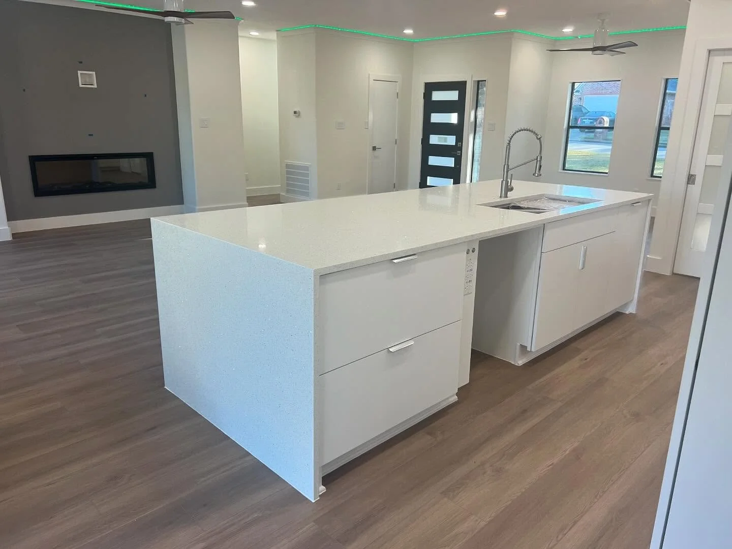 Modern kitchen with a large white island, stainless steel sink, and wooden flooring, opening into a living area with gray accent wall and a gas fireplace.