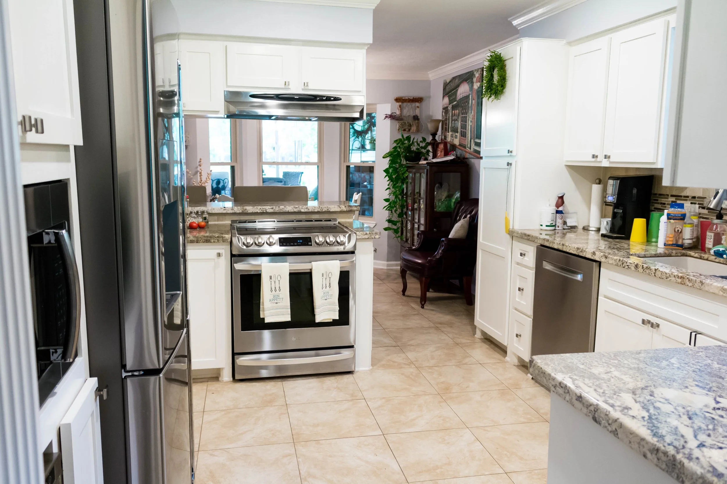 A clean kitchen with white cabinets, stainless steel appliances, and granite countertops. There is a stove with two towels hanging on the oven handle, a refrigerator, and a dishwasher. The kitchen opens into a dining area with a table and chairs, vis