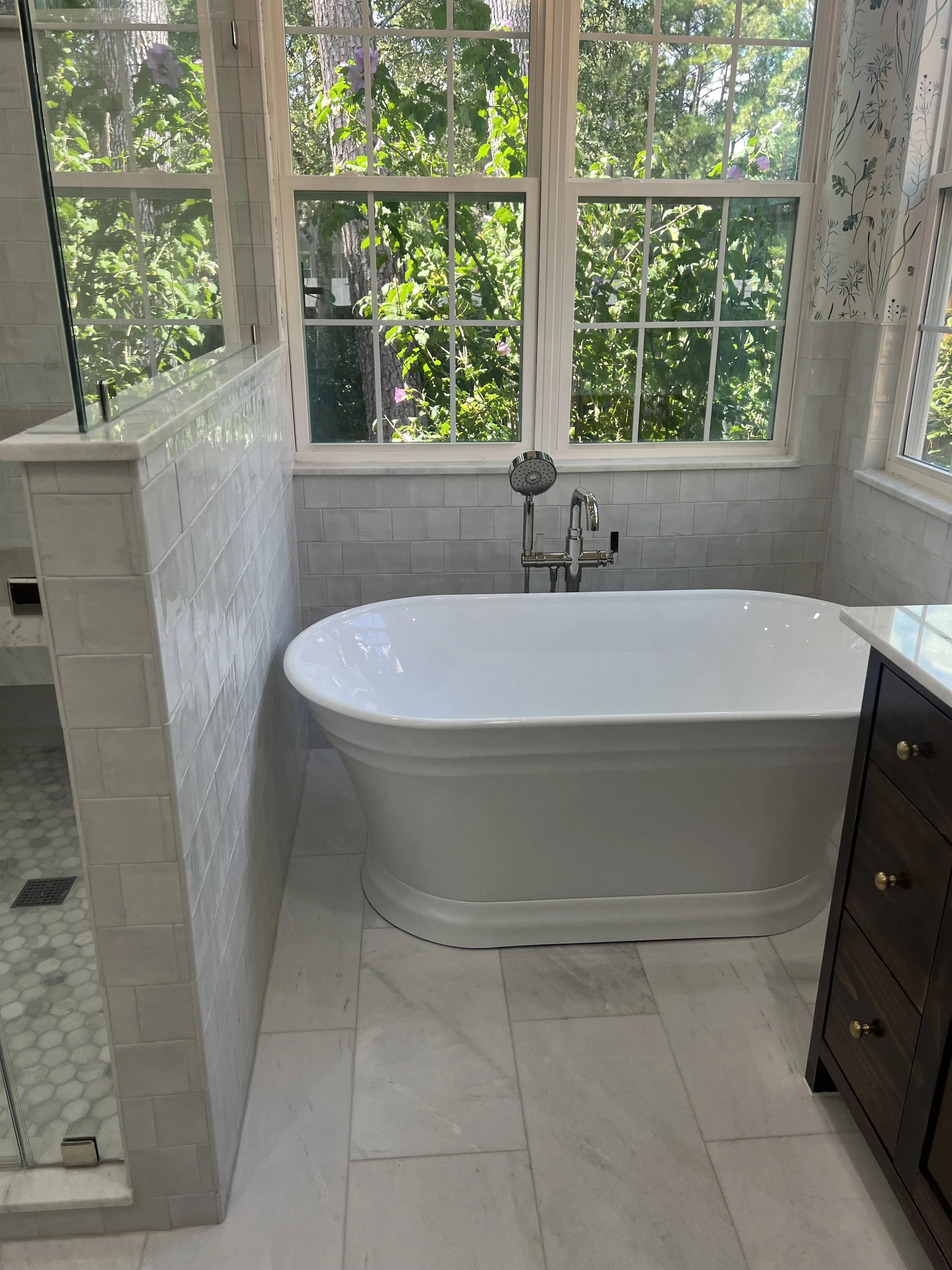 A bathroom with a freestanding bathtub positioned in front of a large window that overlooks lush greenery outside. The window features multiple panes and a white frame. To the left, there is a glass shower with a small tiled shower area, and to the r