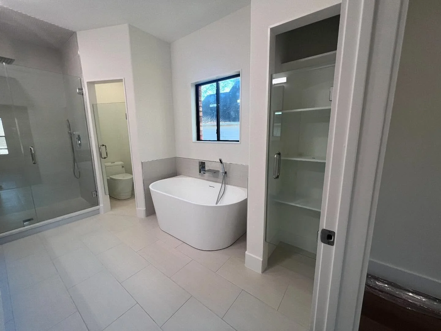 Modern bathroom with glass-enclosed shower, toilet, freestanding bathtub, window, and open shelves for storage.