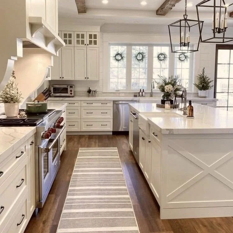 Bright kitchen with white cabinets, a large island, stainless steel appliances, and a window with four wreaths. Decor includes plants, a vase, and candles.