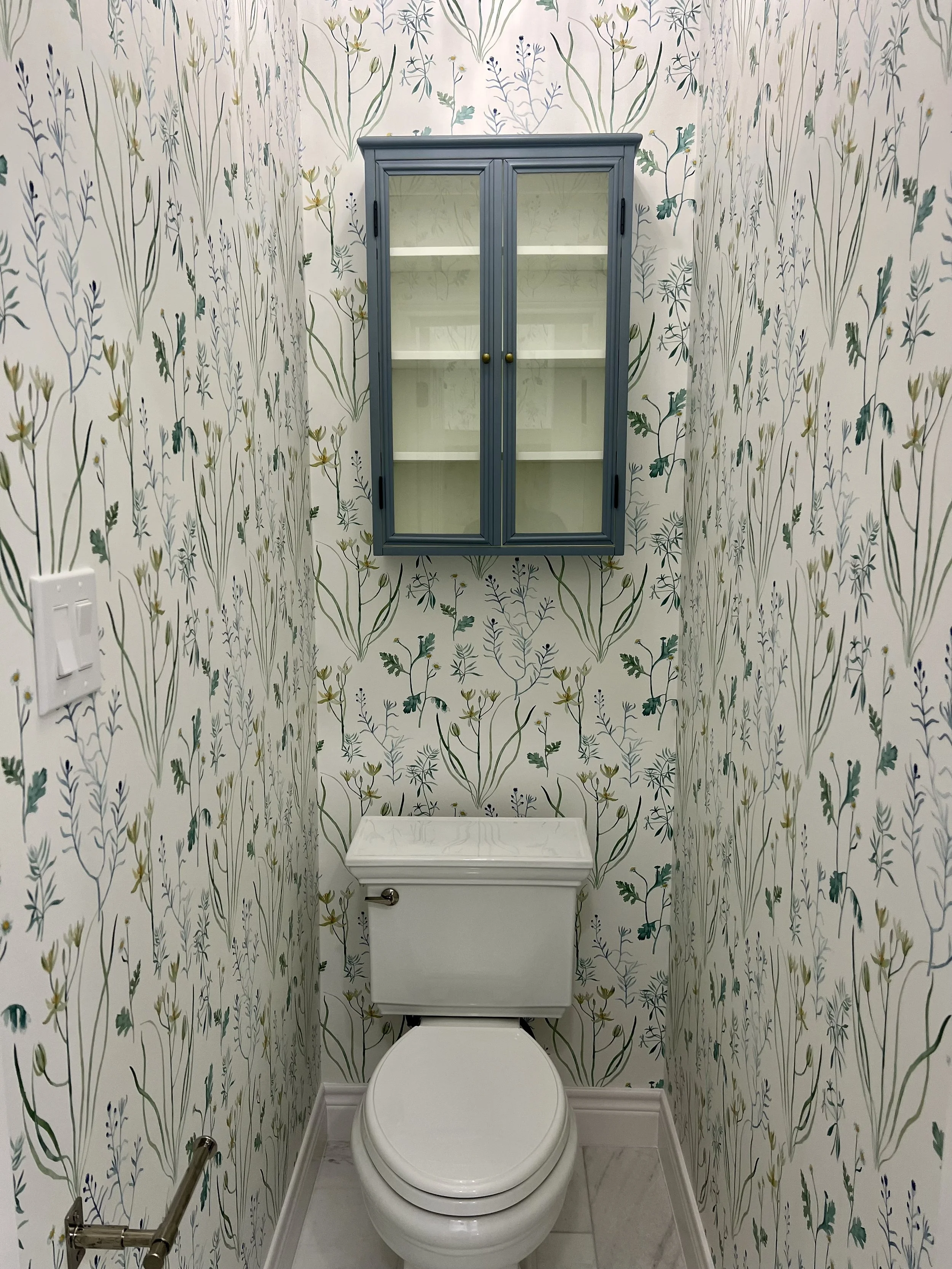 A small bathroom with floral patterned wallpaper, a white toilet, a blue wall-mounted cabinet with glass doors, a white shelf inside, a wall-mounted toilet paper holder, and a flush button on the wall.