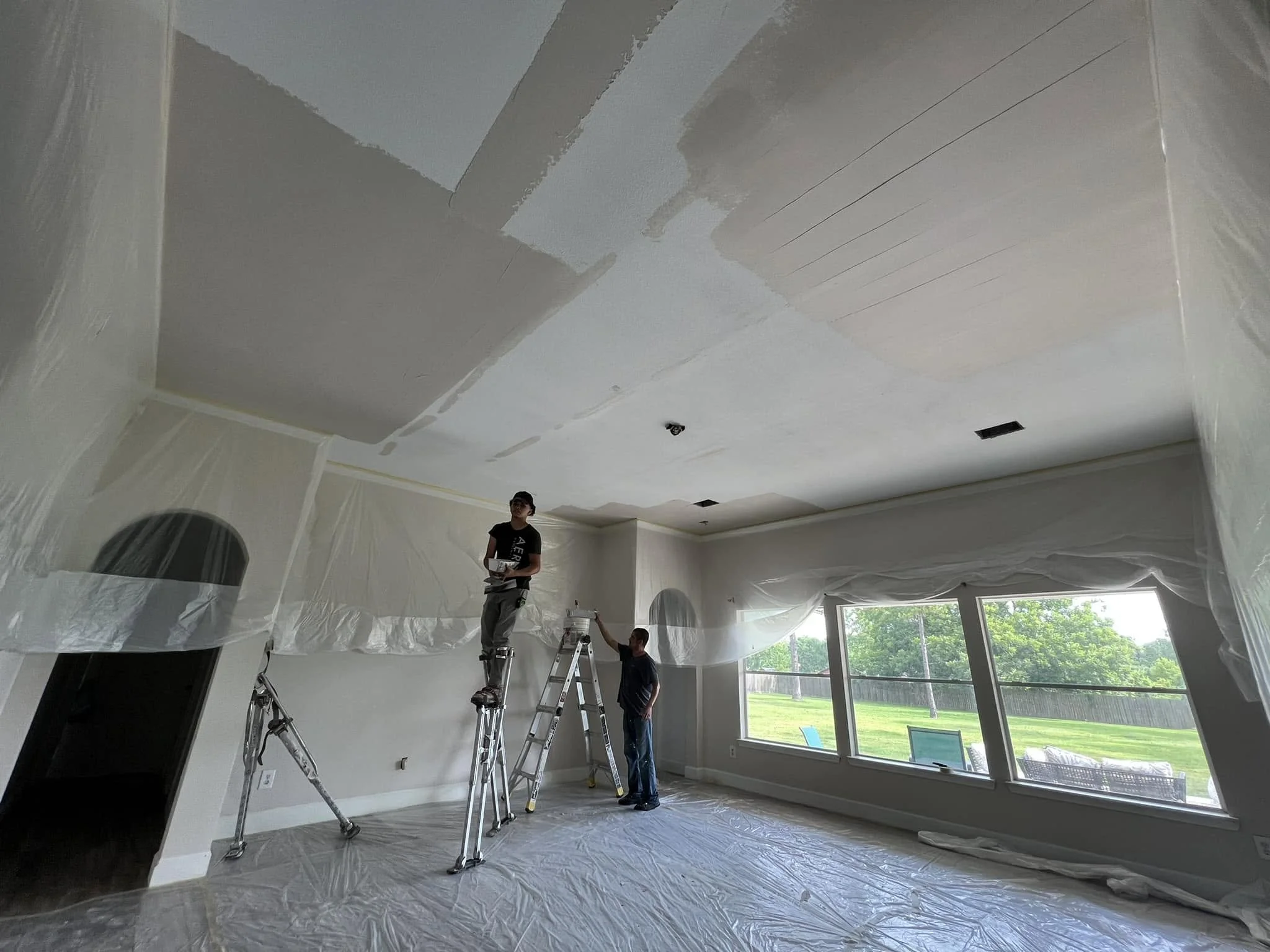 Two workers painting a ceiling in a living room. One is on a ladder, and the other is standing on the floor. The room has large windows and is covered with plastic sheeting to protect surfaces during painting.