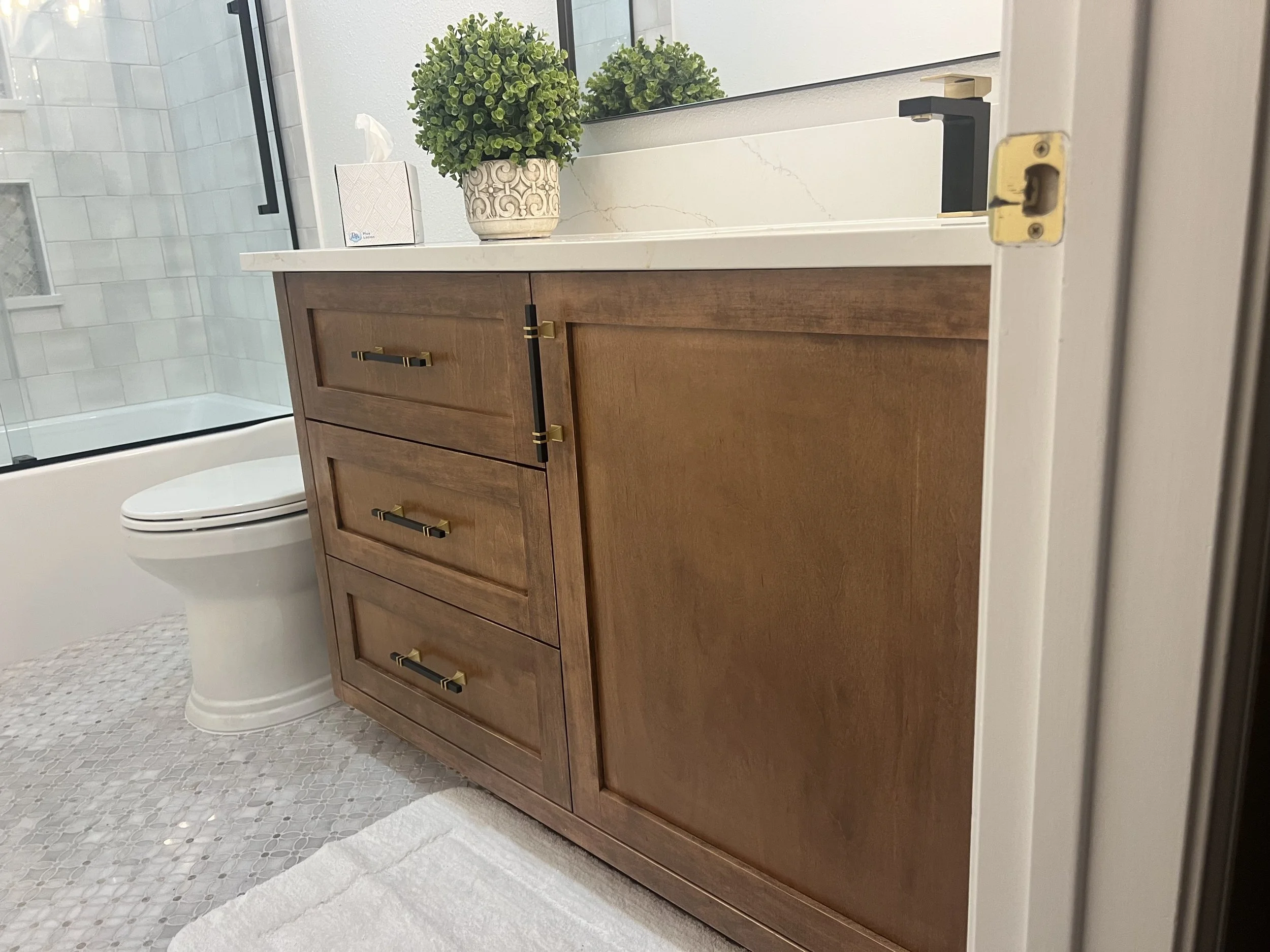 View of a bathroom vanity with a wooden cabinet, a white marble countertop, and a black faucet. On the countertop, there is a potted plant with green foliage and decorative pot, and a box of tissues. To the left, part of a white toilet and a glass sh