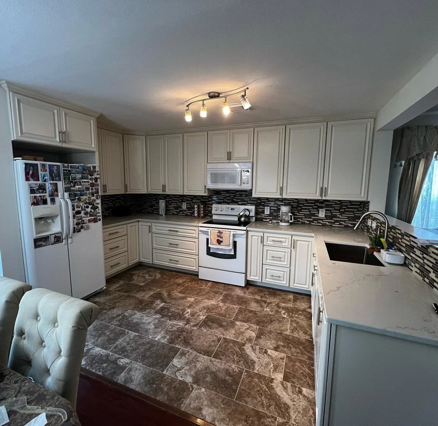 Kitchen with white cabinets, black and gray backsplash, white stove, microwave, refrigerator with photos, marble countertops, sink, and brown tiled floor.
