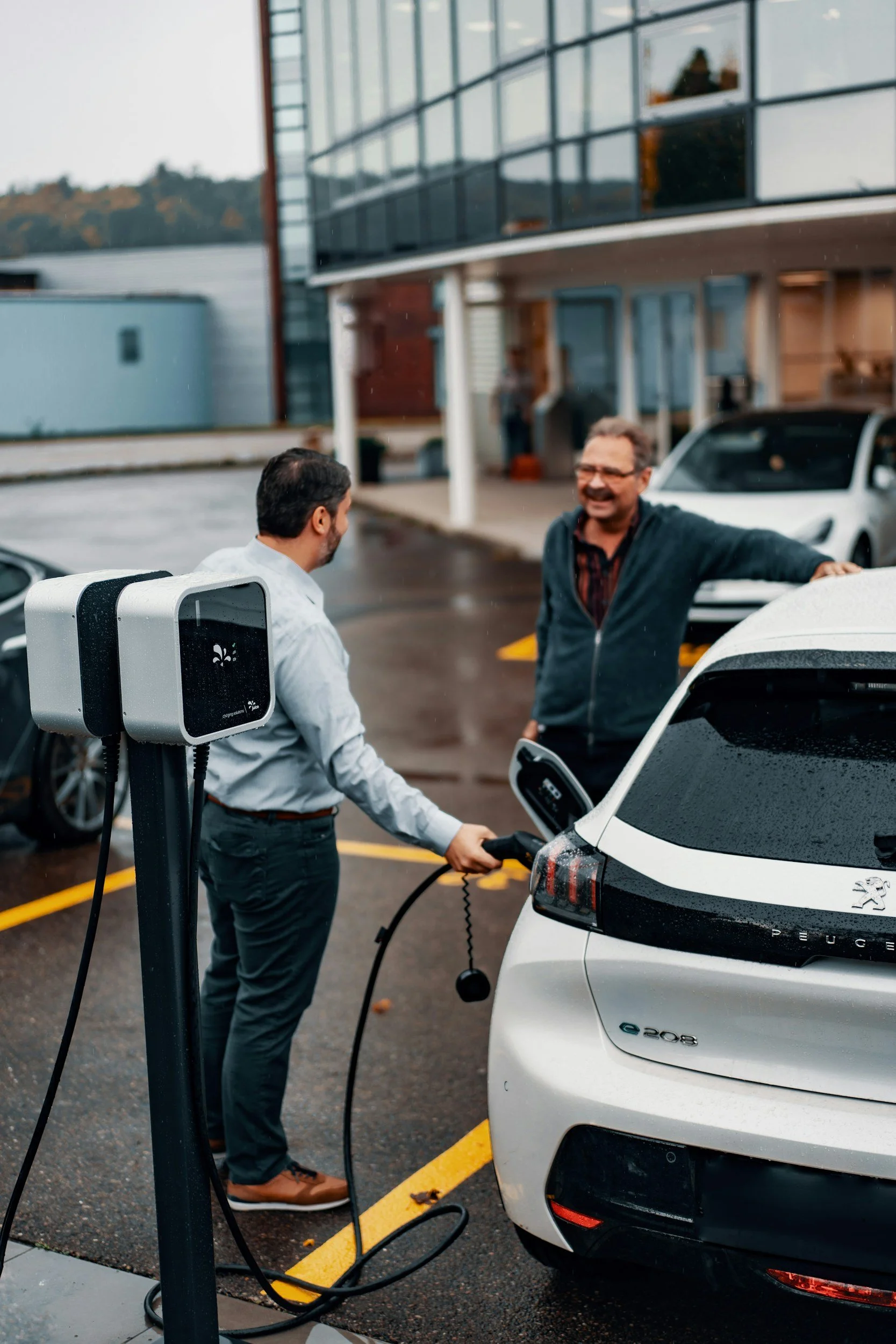 Two men smiling and chatting at an electric vehicle charging station. One man is plugging in a white Peugeot e-208. The charging station has a digital display. The background shows a building with large glass windows and parked cars in a parking lot.