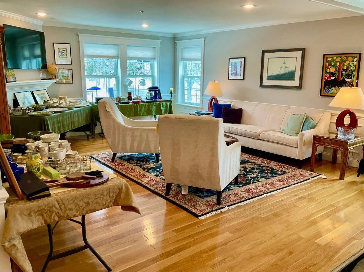 Living room with large windows, white sofa, cream armchair, two table lamps, framed artwork, wooden side table, patterned rug, and various dishes and items on tables, likely set up for a gathering or brunch.