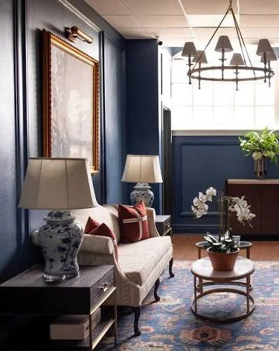 Living room with navy blue walls, a beige sofa with red accent pillows, two table lamps with floral bases, a round coffee table with a potted orchid, large window with grid pattern, and a chandelier hanging from the ceiling.
