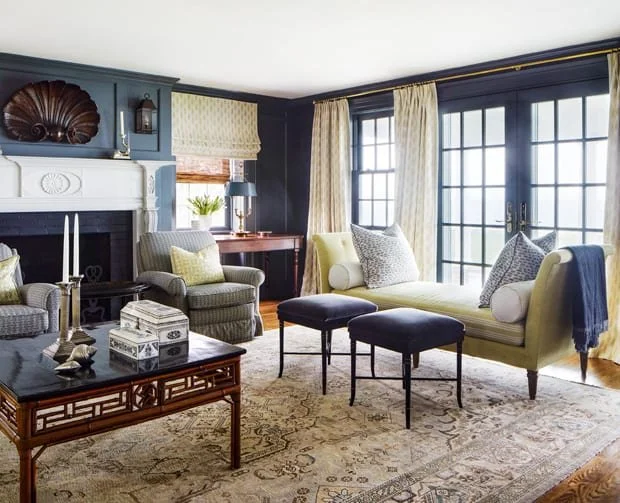 Living room with navy blue walls, large windows with cream curtains, a white fireplace, a yellow sofa with throw pillows, two black ottomans, a patterned armchair, a wooden coffee table, and a window seat with cushions.