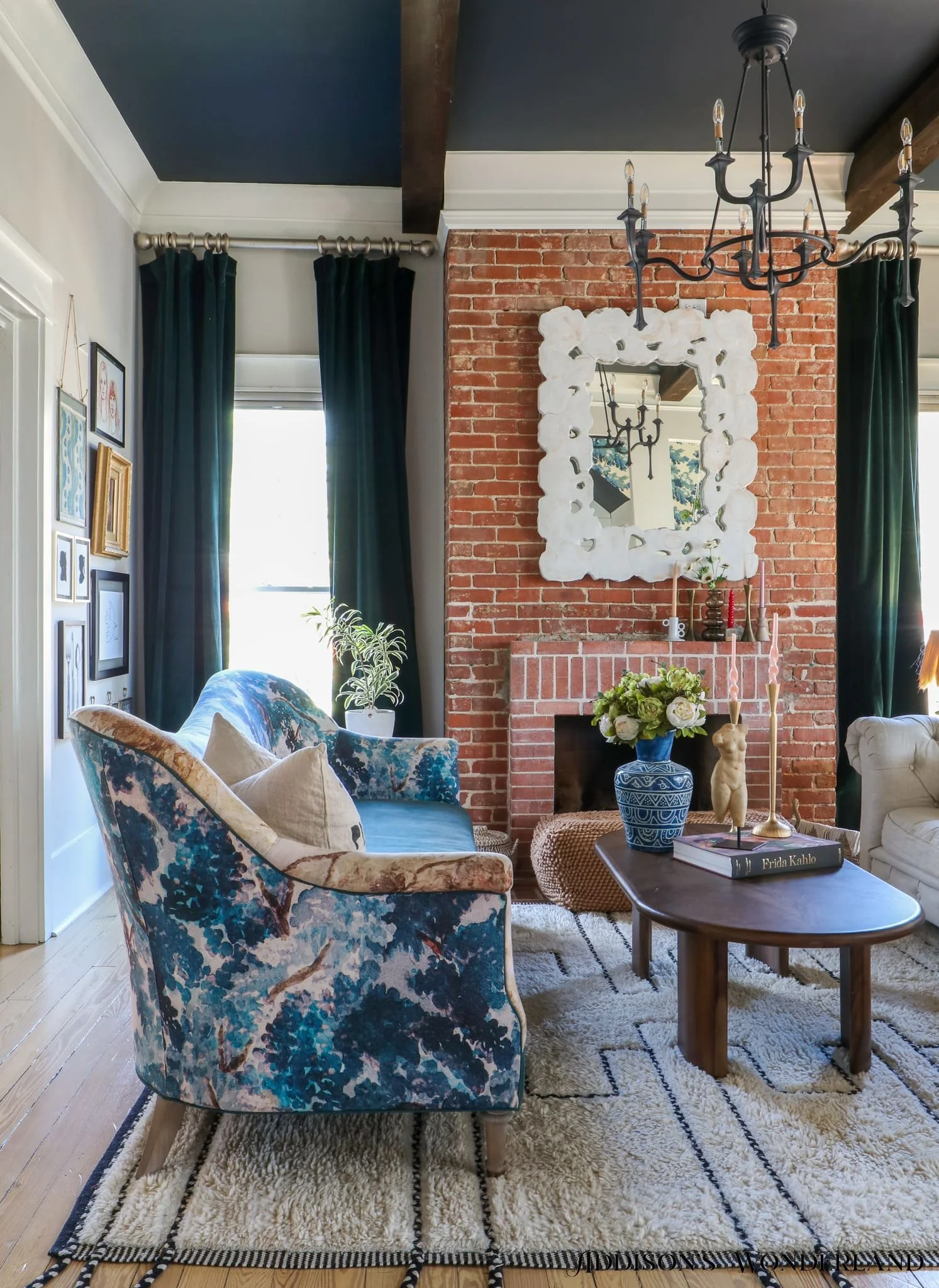 A living room with a brick fireplace, a black chandelier, and large windows with dark green curtains. There is a floral-patterned armchair, a coffee table with books and decor, and artwork on the wall.