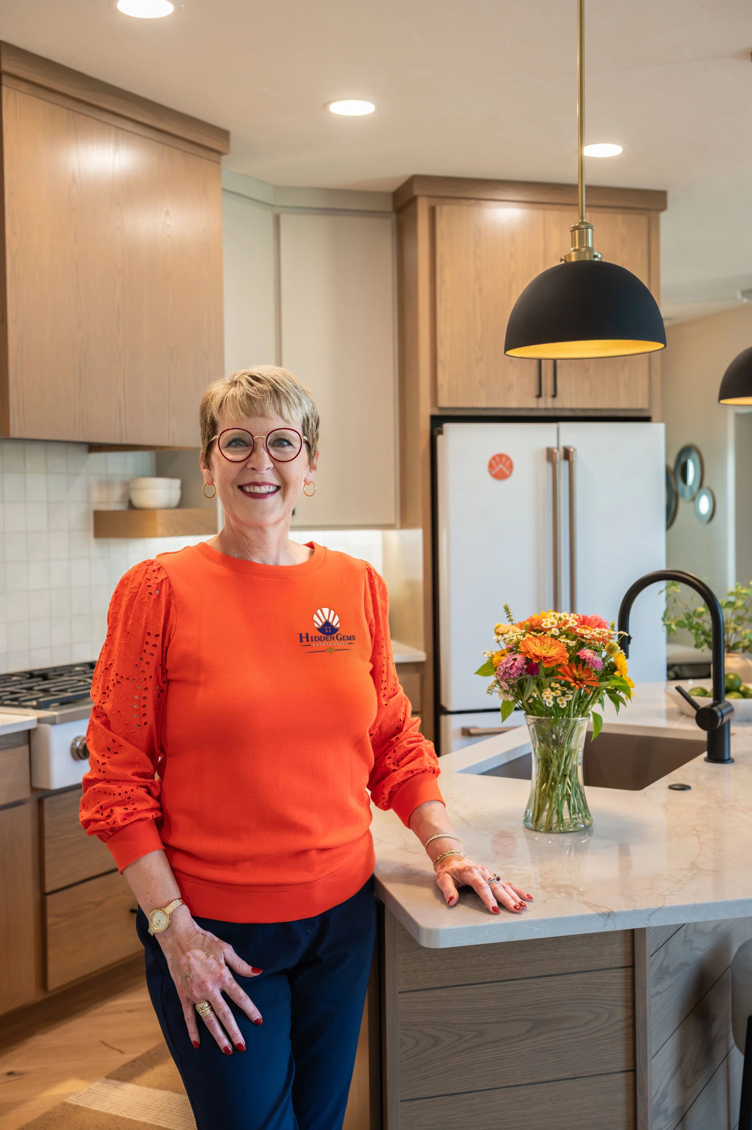 A woman with short blonde hair and glasses stands in a modern kitchen, smiling at the camera. She wears an orange top with eyelet sleeves and dark pants. There is a bouquet of colorful flowers on the cream-colored marble kitchen island, and the background features wooden cabinets, a white refrigerator, and black pendant lighting.
