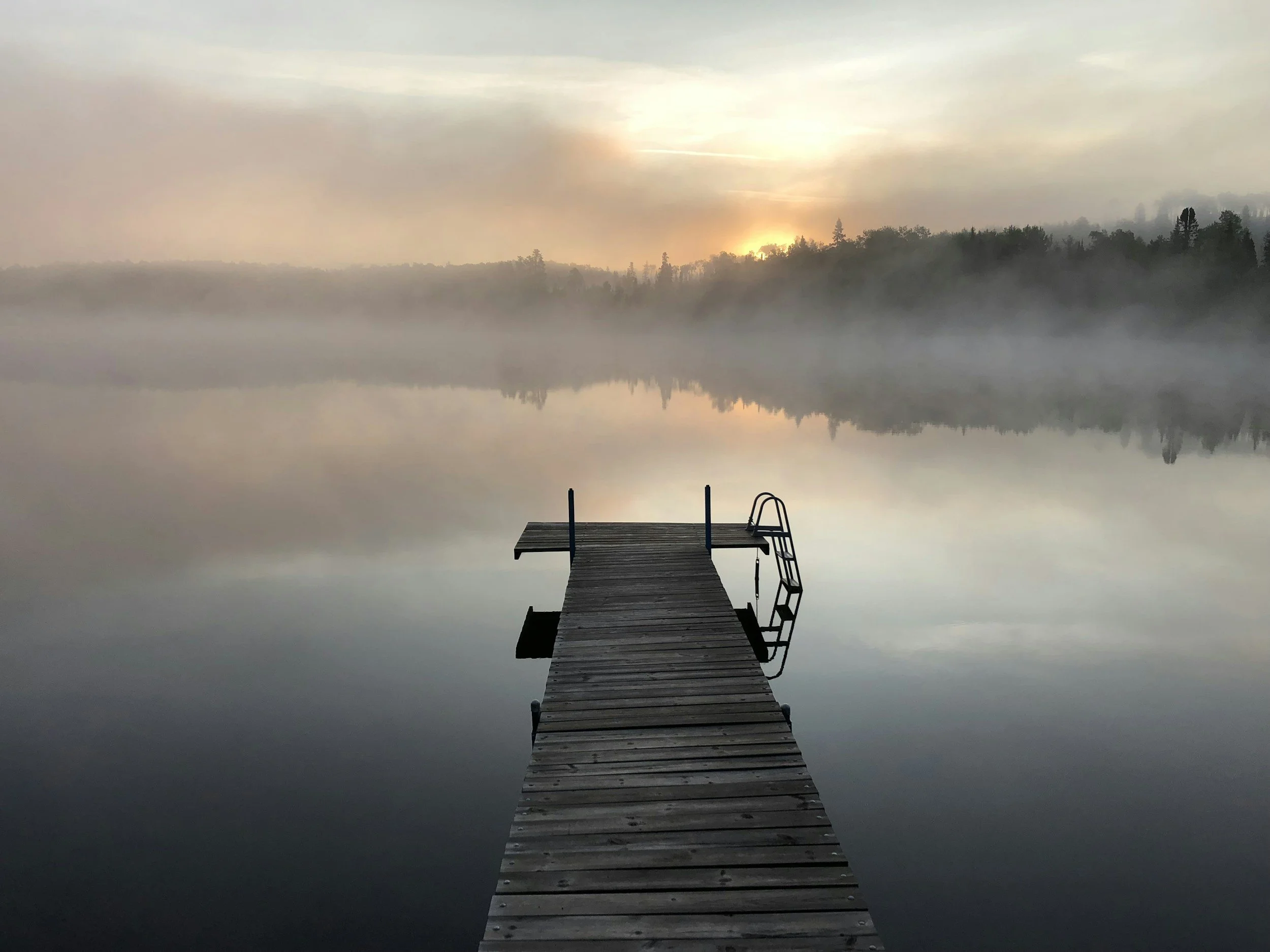 A wooden dock extending into a calm, foggy lake with a ladder at the end. The fog is thick on the water, and the sunrise or sunset is visible through the clouds, casting a soft glow over the scene.