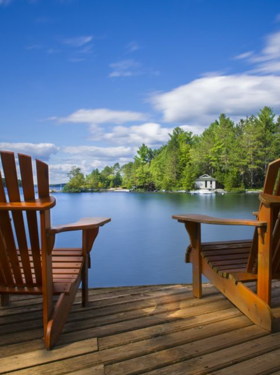 Two wooden Adirondack chairs on a dock overlooking a tranquil lake, with trees and a small white house on the opposite shore under a partly cloudy blue sky.