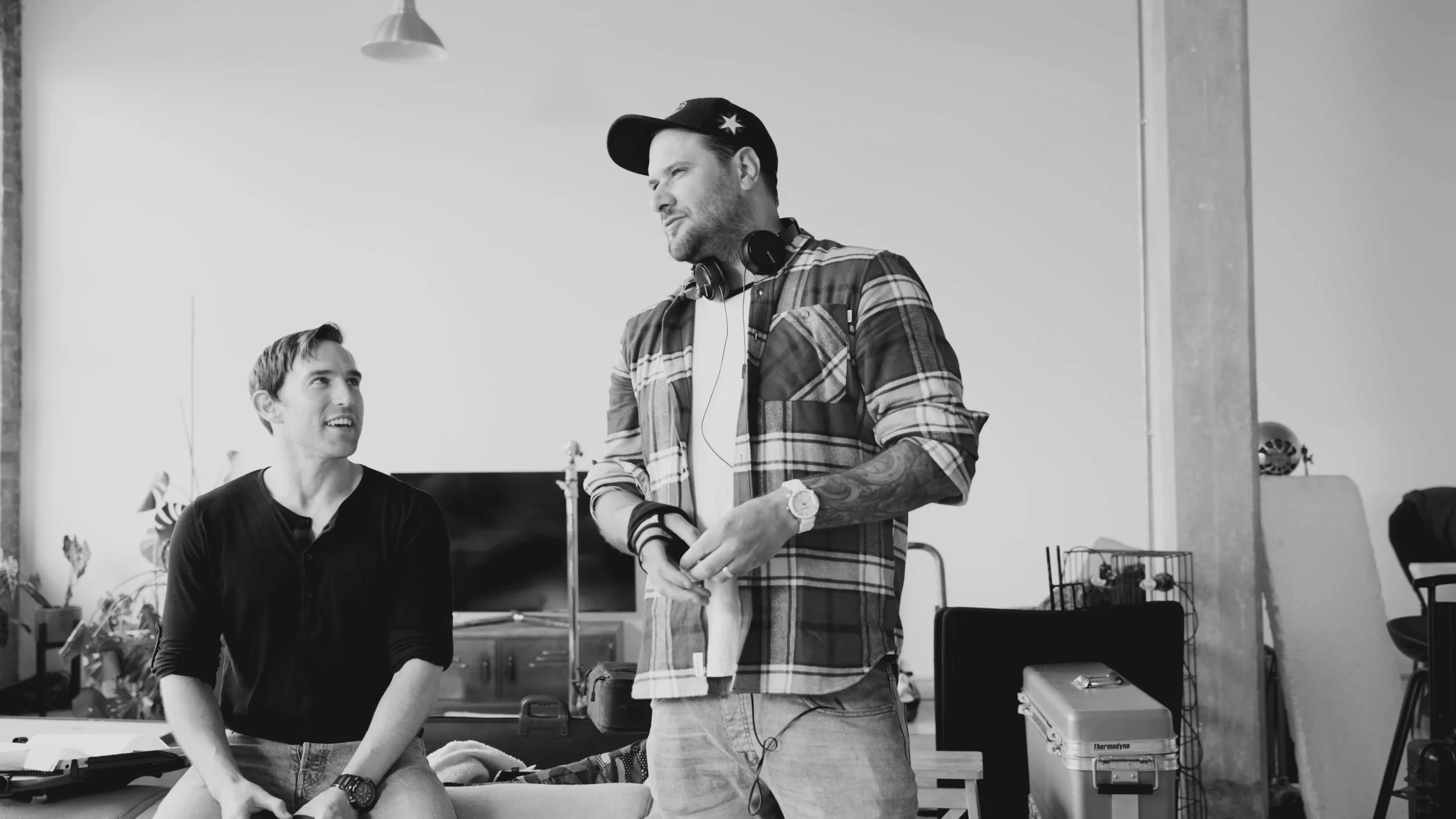 Two men talking in an indoor setting. One man is sitting and looking up at the other man who is standing. The standing man has tattoos on his arm, wears a plaid shirt, a watch, and has headphones around his neck.