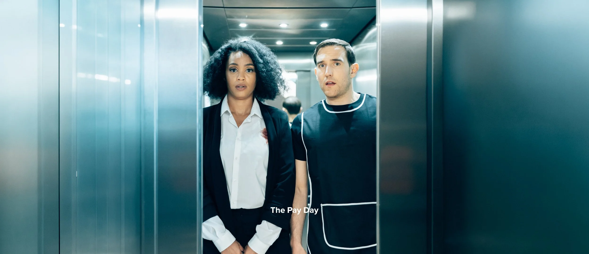 A woman and a man standing inside an elevator, looking surprised or confused.
