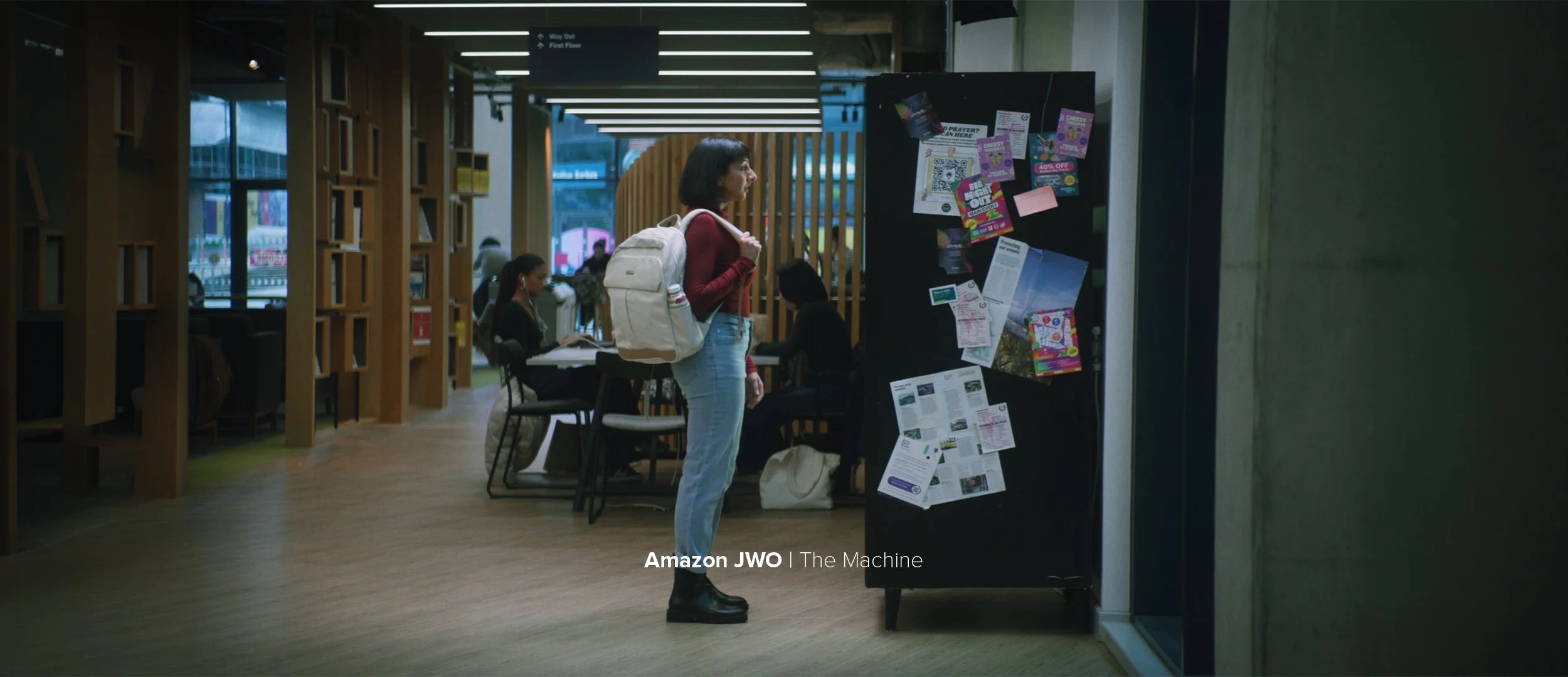 A woman with a cream-colored backpack standing in front of a black bulletin board with colorful flyers and notices in a modern, well-lit public space, possibly a library or community center.