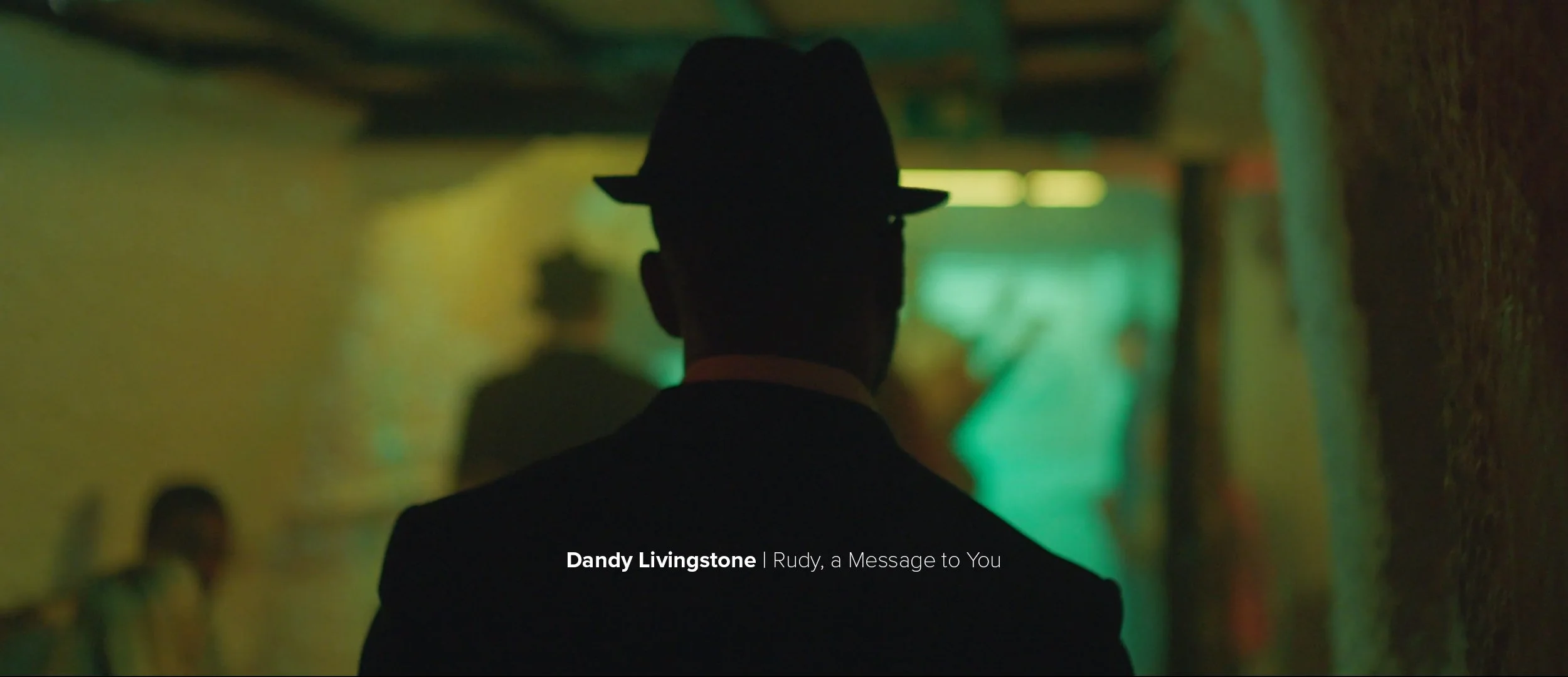 Silhouette of a man wearing a suit and fedora hat, walking in a dimly lit alleyway with green and yellow lighting, with text overlay reading 'Dandy Livingstone | Rudy, a Message to You'.