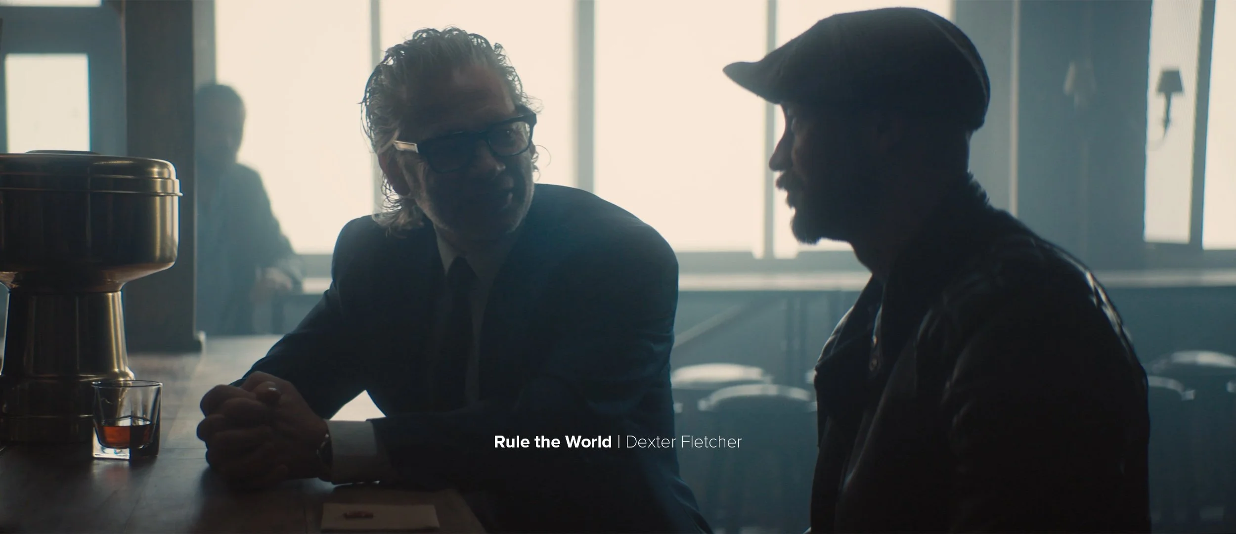 Two men sitting at a bar or diner table having a conversation, with a large window in the background. One man has curly hair and glasses, the other wears a cap and leather jacket. A glass of drink and a large bowl are on the table.