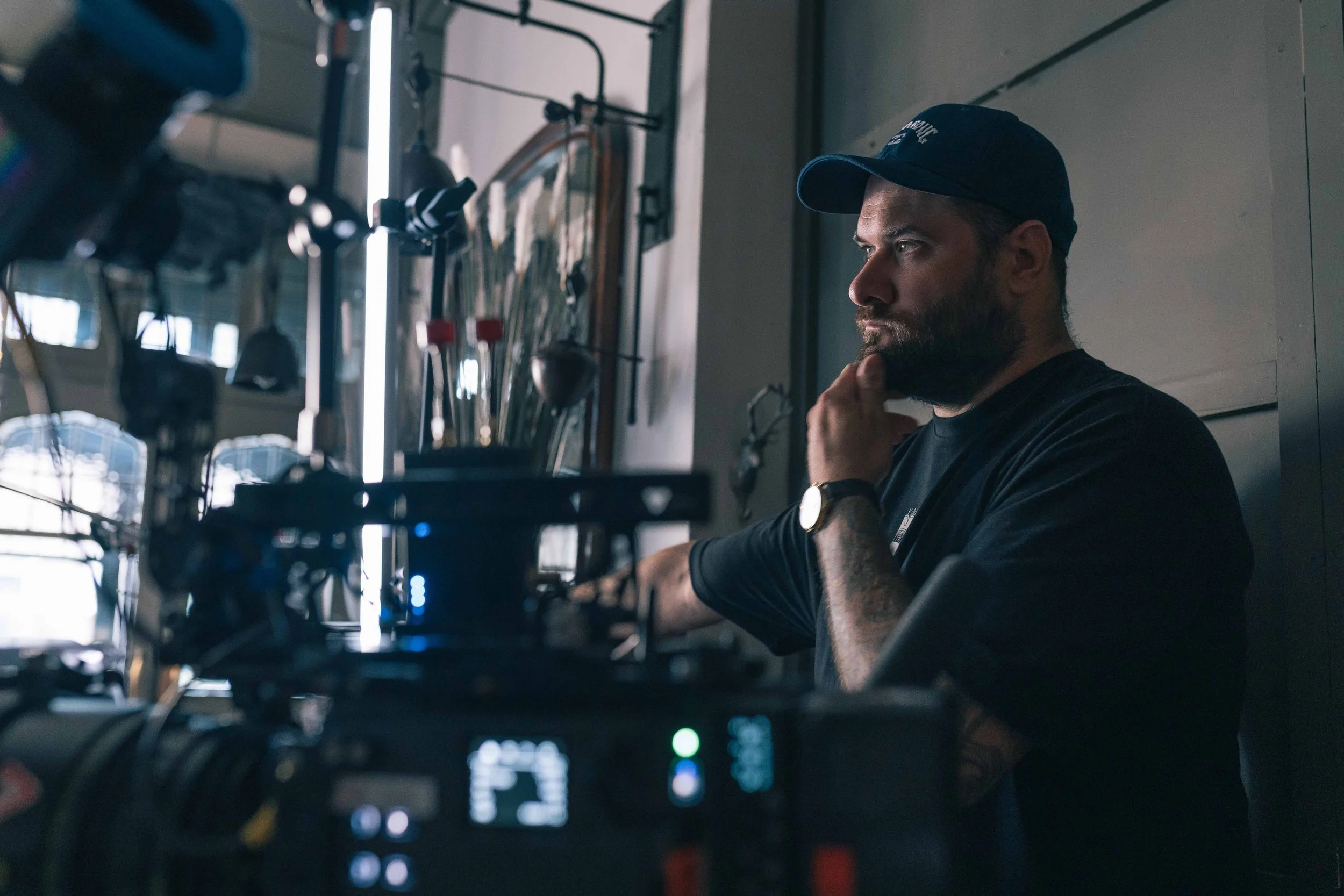 A bearded man wearing a blue baseball cap and black shirt, sitting in a room with various tools and equipment on the wall behind him, appears to be in a workspace or workshop.