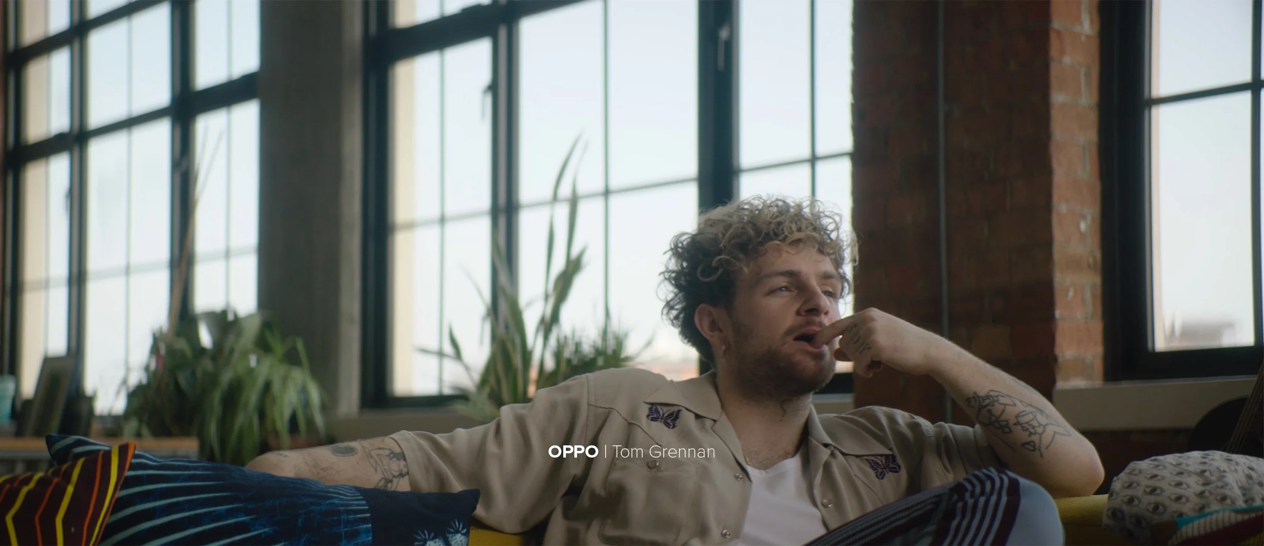 A young man with curly hair and tattoos is sitting on a sofa, resting his chin on his finger, in a room with large windows and a brick wall, with plants in the background.