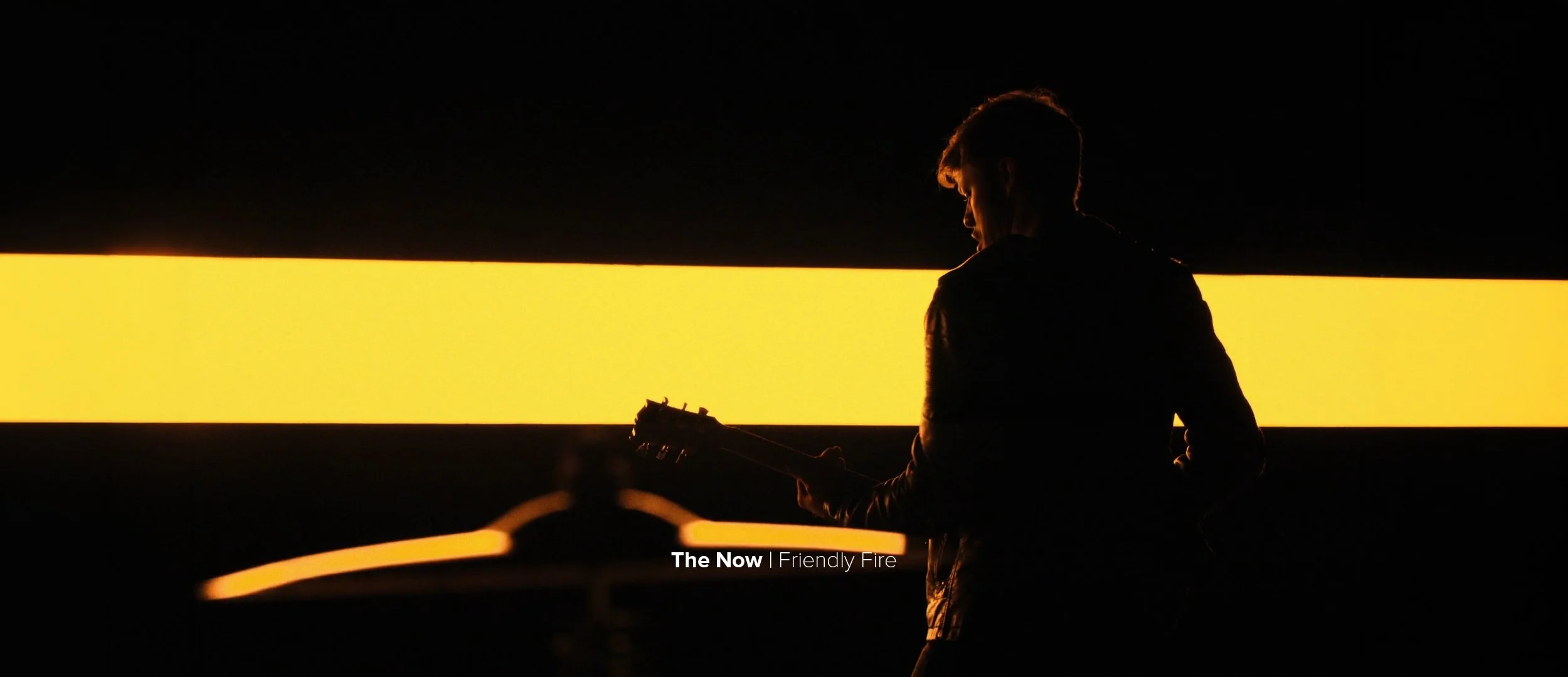 Silhouette of a young man playing an electric guitar in front of a glowing yellow horizontal panel at night.