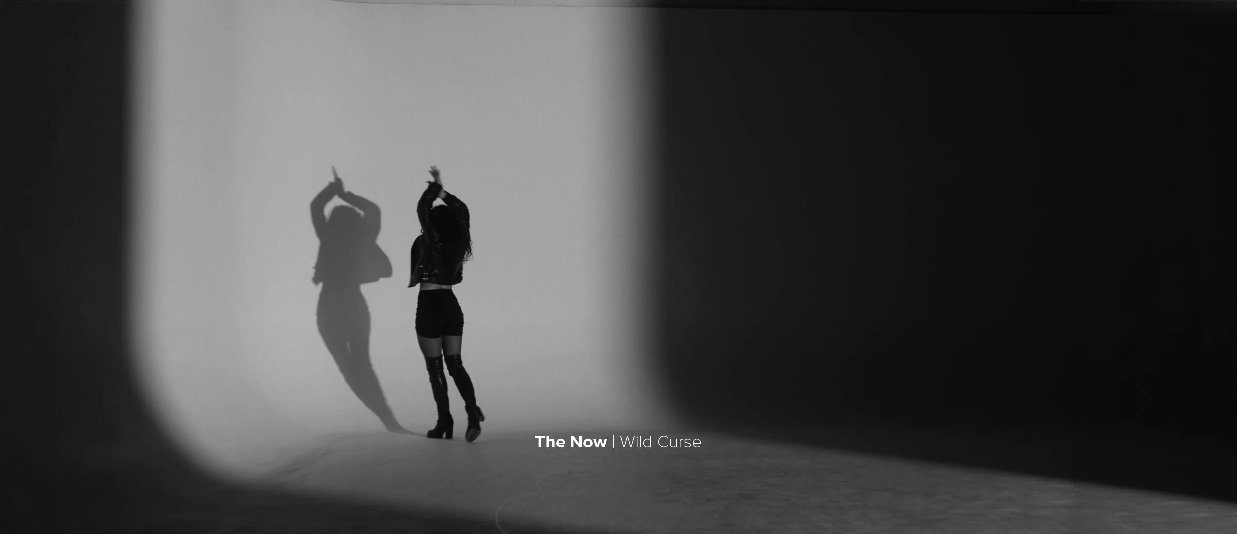 A woman standing against a light background, with her shadow cast on the wall, raising her arms above her head. The scene is in black and white and has a moody, artistic feel.