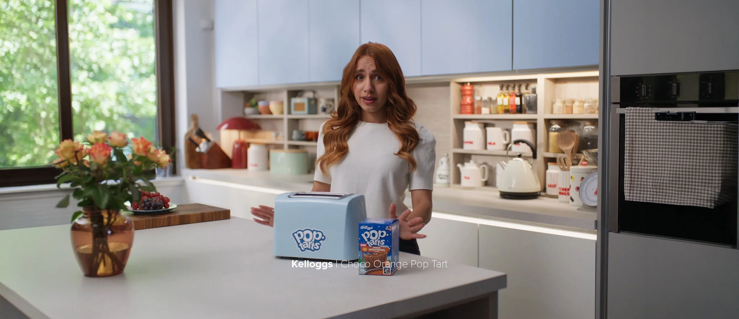 A woman standing in a modern kitchen with a surprised or confused expression, near a box of Kellogg's Choco Orange Pop Tarts on a white kitchen counter with a flower arrangement, in front of a window with green trees outside.