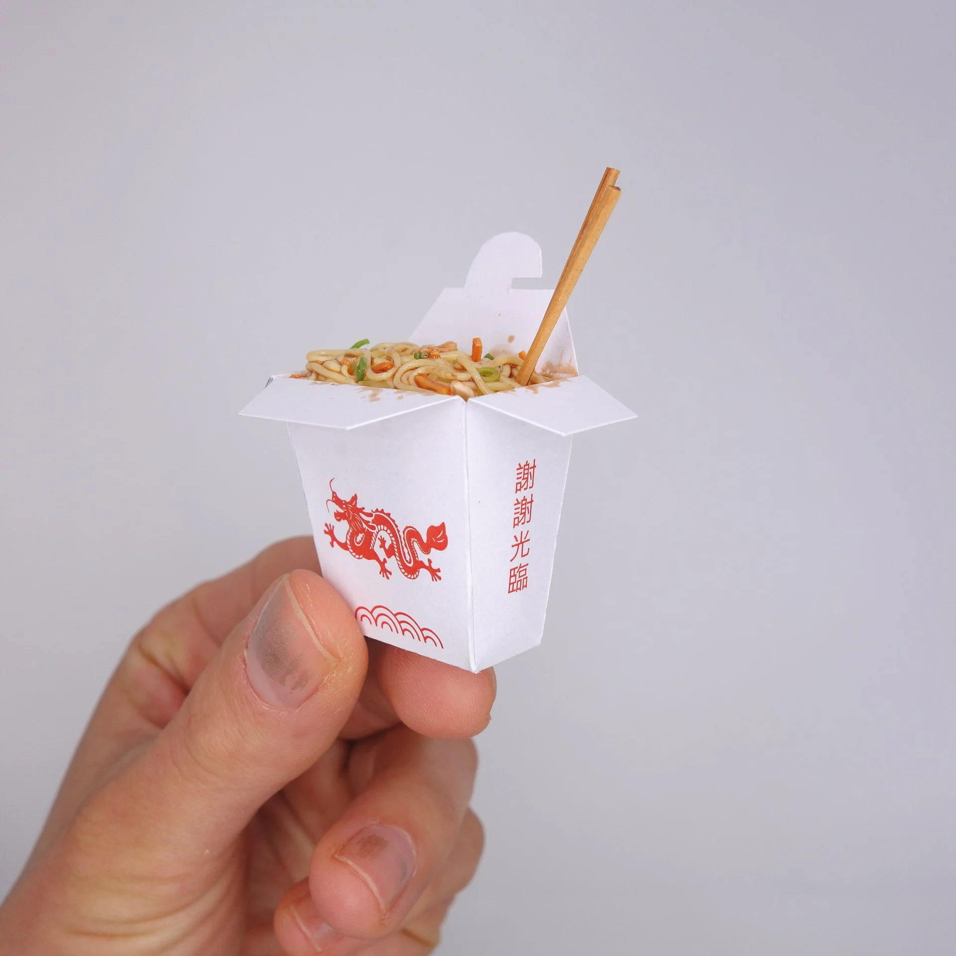 Nahaufnahme einer Hand, die eine chinesische Takeaway-Box mit gebratenen Nudeln hält, vor einem neutralen grauen Hintergrund.
