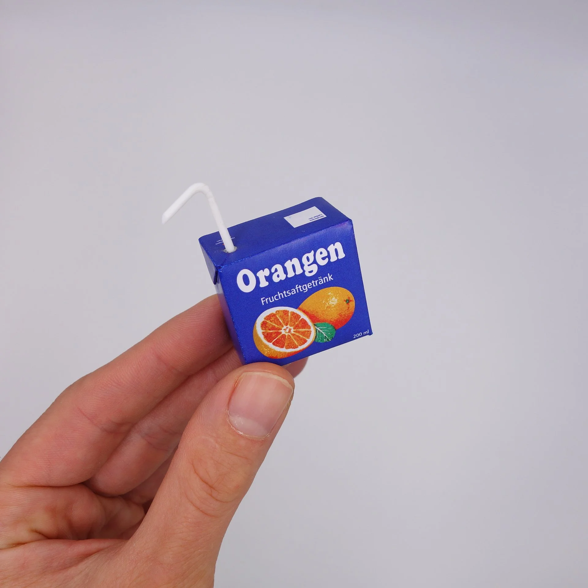 Nahaufnahme einer Hand, die eine kleine blaue Tetra Pak Verpackung mit_orangenem Inhalt hält. Die Verpackung hat einen orangen Ring und zeigt eine Orange und einen Limettenzuschnitt, sowie den Text "Orangen" und "Fruchtsaftgetränk". Der Strohhalm ragt aus der Verpackung heraus.