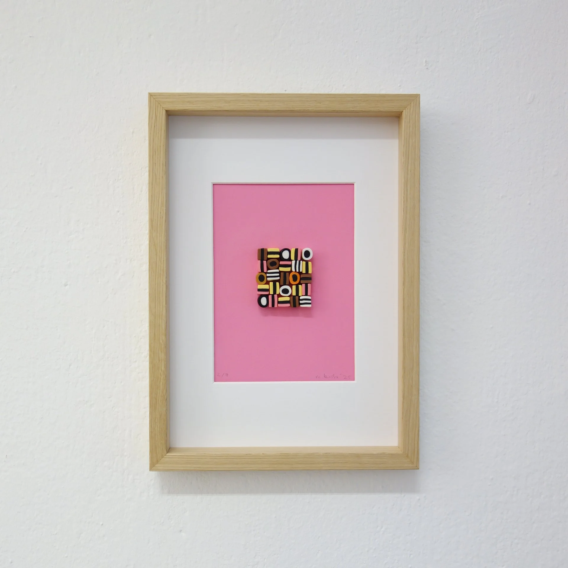 Kunstwerk mit schwarzmusterartigen Formen auf rosa Hintergrund, gerahmt in Holz, an weißer Wand.
