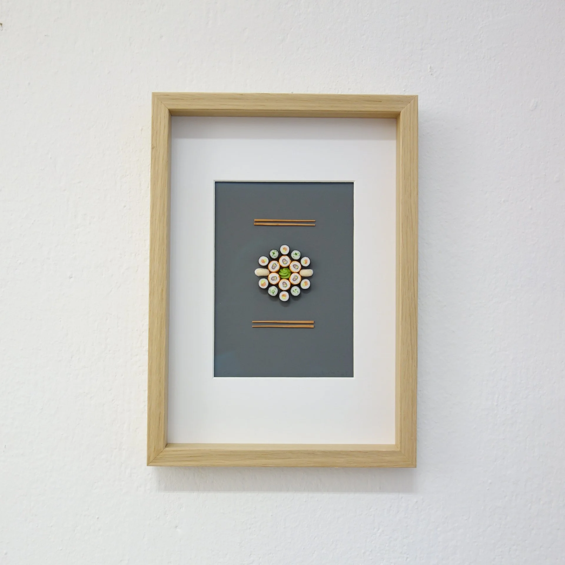 Schwarze Pinnwand mit einem runden Arrangement aus kleinen dekorativen Objekten, die die Sushi Zutaten darstellen, in einem Holzrahmen an einer weißen Wand.