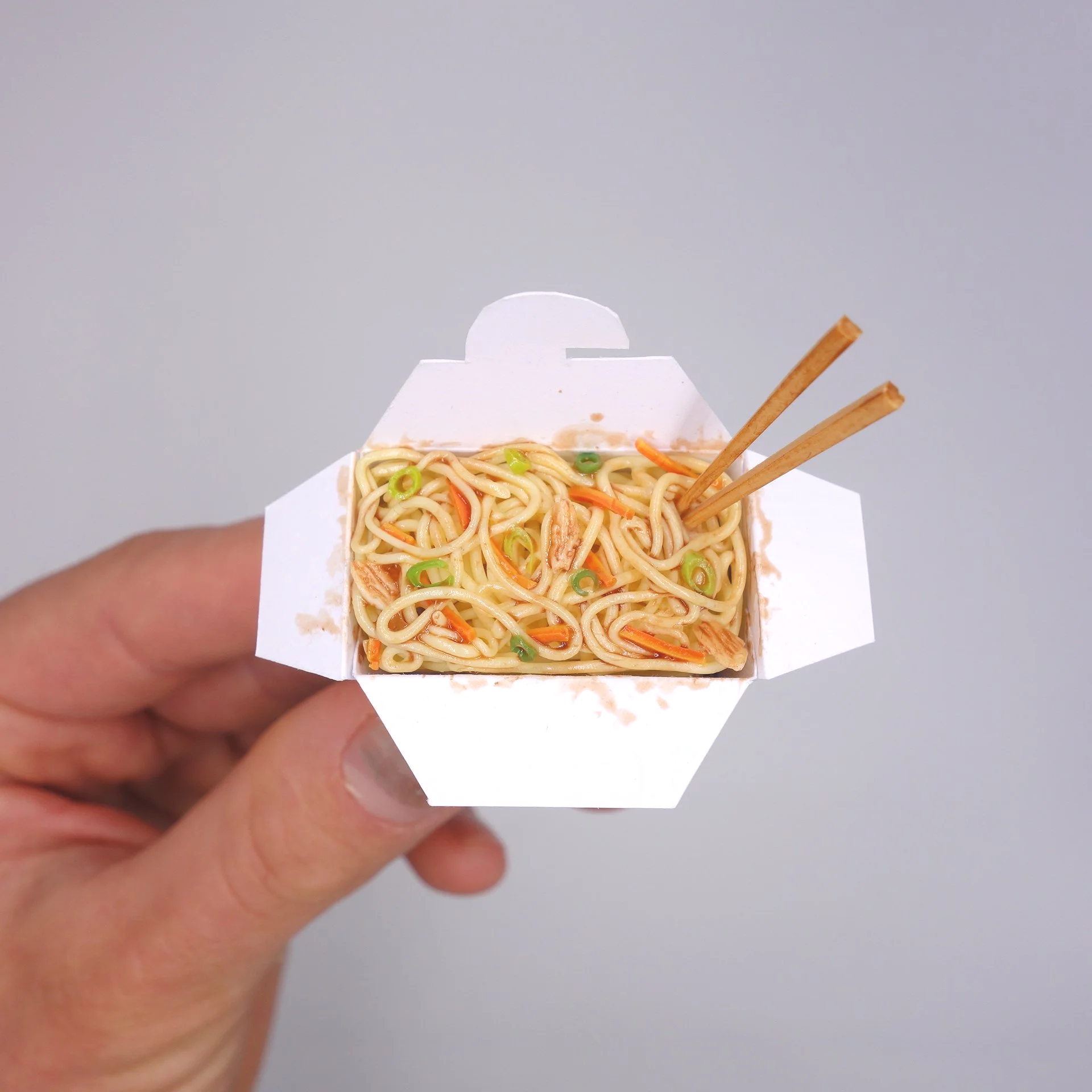 Nahaufnahme eines Takeaway-Containers mit Nudelgericht, in einer Hand gehalten, mit Essstäbchen auf der rechten Seite.