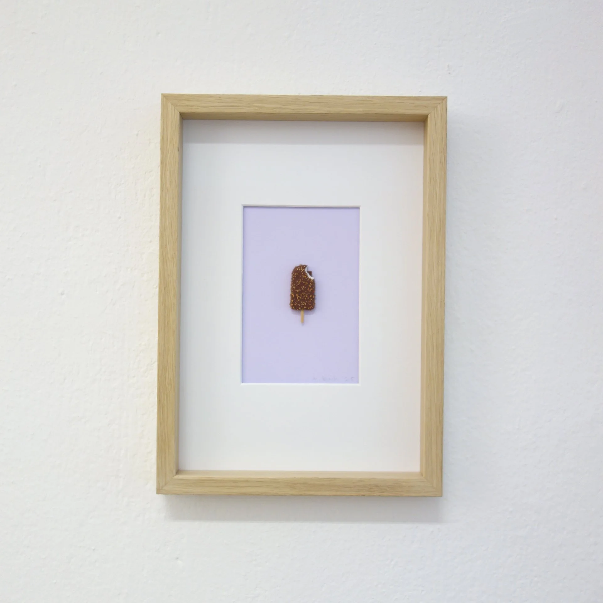Ein Bilderrahmen hängt an einer weißen Wand und enthält ein Foto eines Schokoriegels auf einem Holzstäbchen.