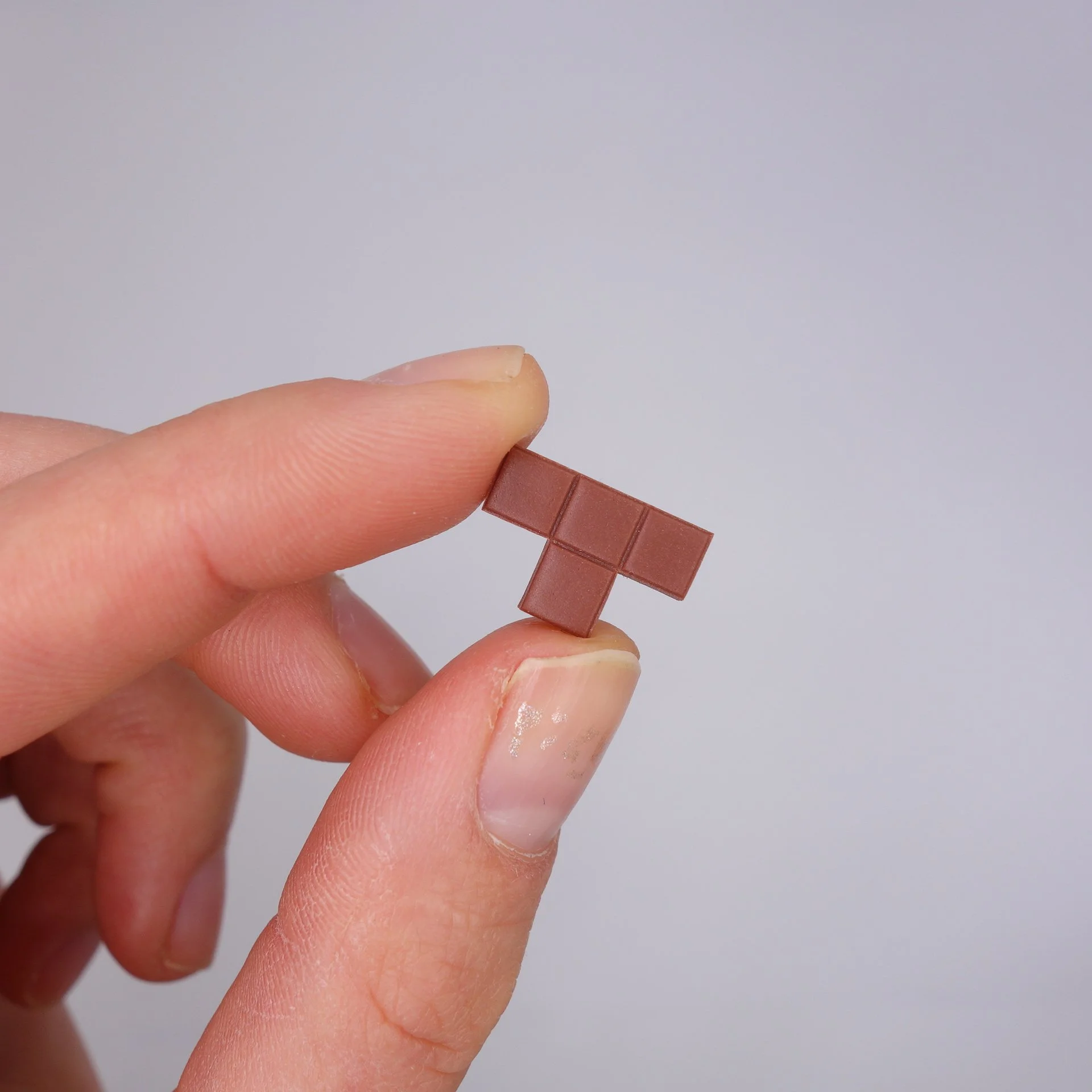 Nahaufnahme einer Hand, die einen kleinen Brick-Schokoladenriegel zwischen Daumen und Zeigefinger hält, vor hellem, neutralem Hintergrund.